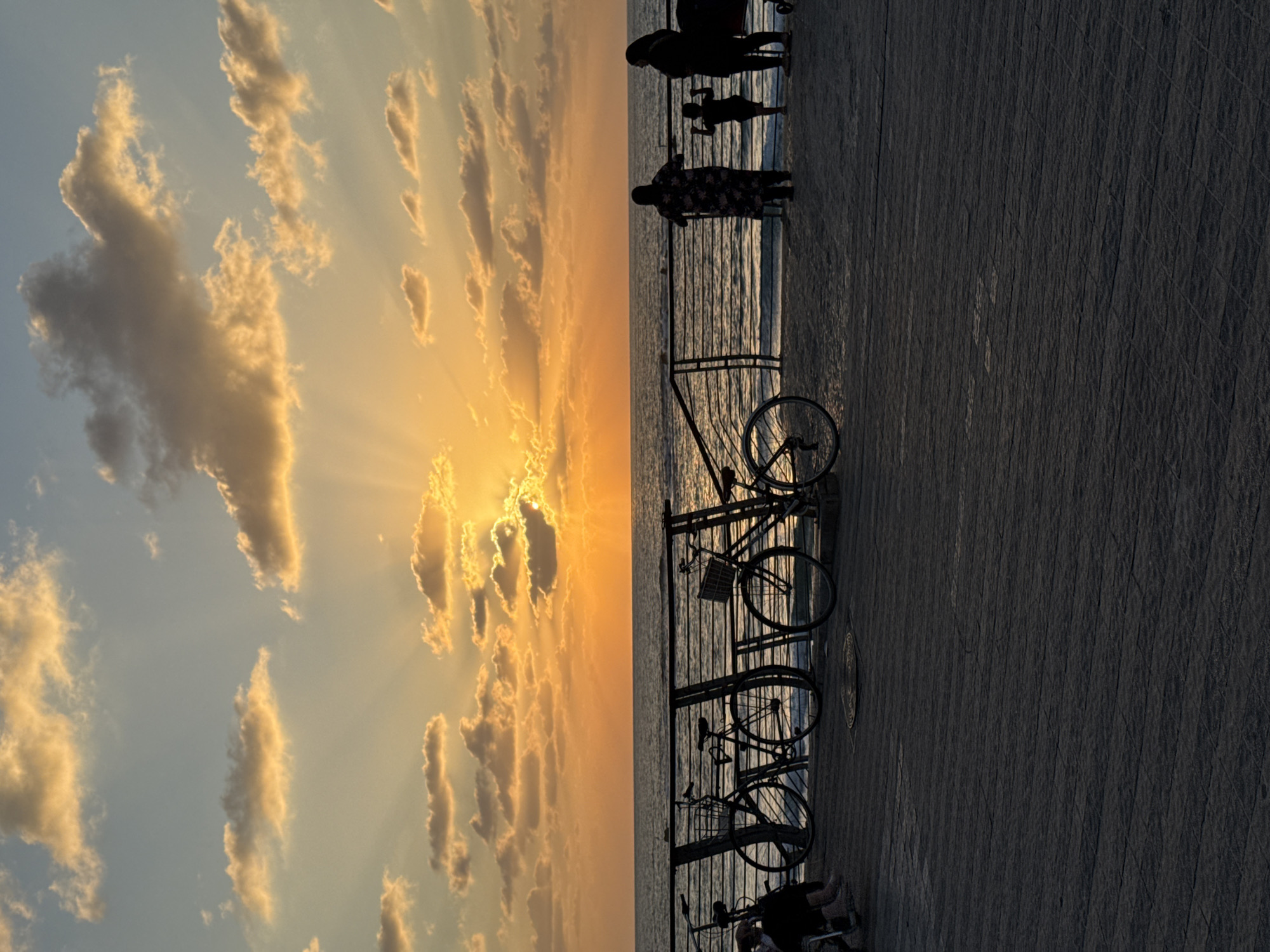 This image captures a serene sunset over the Mediterranean Sea, likely from the Tel Aviv promenade, also known as Tayelet. Silhouettes of people and bicycles dot the boardwalk, which is paved with what appears to be dark stone tiles. A metal railing separates the boardwalk from the sea, where gentle waves lap against the shore. The sky is a canvas of warm oranges and yellows, with dramatic sunbeams piercing through scattered clouds. Several bicycles are parked along the railing, suggesting a leisurely evening stroll or bike ride. In the foreground, a few individuals, including a child, are visible, their figures outlined against the vibrant sunset. The overall atmosphere is peaceful and contemplative, a common scene enjoyed by locals and tourists alike on Tel Aviv's coastline.
