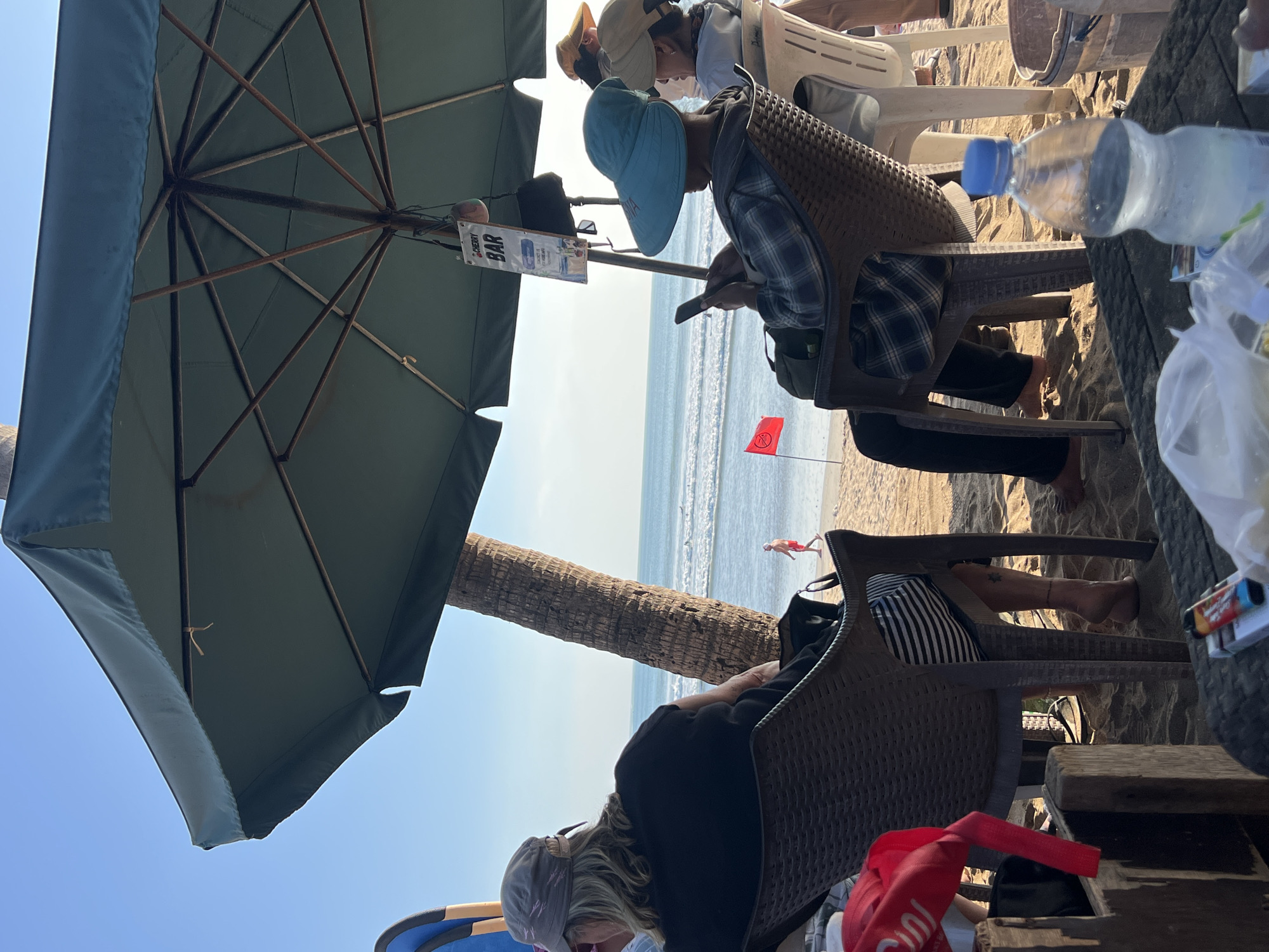 In Kuta, Indonesia, under a large teal umbrella on a sandy beach, two individuals are seated in chairs, facing the ocean. The person on the right, wearing a wide-brimmed blue hat and a plaid shirt, is looking at their phone. To their left, another individual, partially visible, is wearing a black shirt and a grey cap. Behind them, a person in a white shirt is seated. In the distance, the ocean has gentle waves, and a man in red shorts walks along the shoreline. A red flag is planted in the sand near the water. A palm tree stands tall next to the umbrella. On a table in the foreground, a bottle of water, a pack of cigarettes, and a lighter are visible. A sign hanging from the umbrella reads "CHERRY BAR."