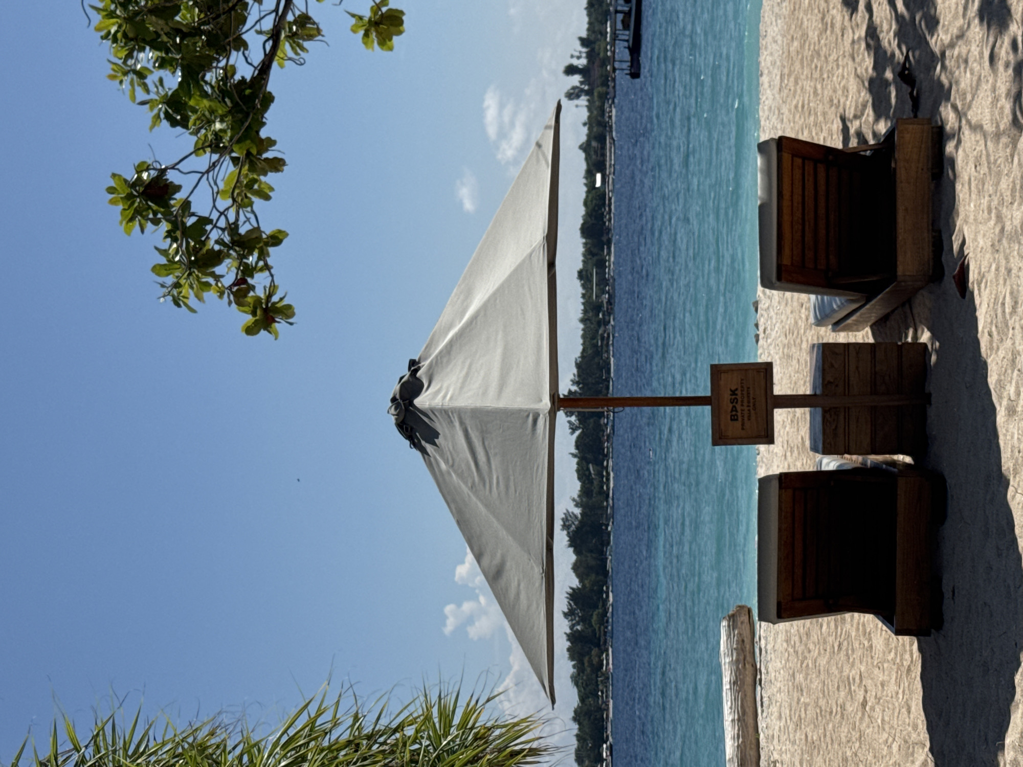 The image depicts a beach scene at an unknown location in Indonesia. The view includes a beach umbrella casting shade over two wooden lounge chairs. The sand appears light in color. Beyond the chairs and umbrella, the turquoise water of the ocean extends towards the horizon, where a strip of land with trees is visible. A sign that reads "BDSK Private Property Villa Residents Only" is positioned next to the lounge chairs. A tree branch with green leaves is visible in the top left corner of the image, while green foliage is visible in the bottom left corner. The sky is blue with some clouds.