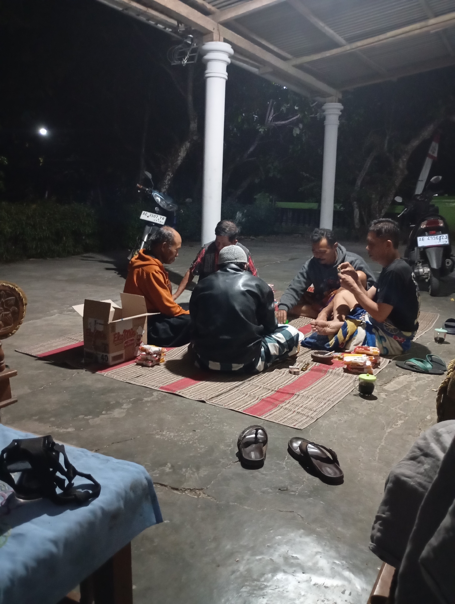 In the city of Wonogondo, Indonesia, a group of five men are gathered on a concrete floor under an open-air shelter at night. They are seated on woven mats. Three men are in the foreground, and two are in the background. The man on the far left is wearing an orange hooded jacket and dark pants. Next to him is a man in a black leather jacket and checkered pants, wearing a white peci hat. To his right, a man in a gray hoodie and patterned sarong is visible. In the background, two more men are seated, one wearing a black t-shirt and sarong.
Several objects are scattered around the group. A cardboard box is open to the left of the man in the orange jacket, containing several small packages. A few cigarette packs are also visible on the mat. To the right of the group, two pairs of flip-flops lie on the ground, one dark brown and one green. A small green cup is near the flip-flops.
Motorcycles are parked in the background, one with a license plate reading "AE 3107 YD" and another with "AE 4736 ZJ." The environment appears to be outdoors, with trees and foliage visible behind the shelter. The lighting suggests it is nighttime, with artificial light illuminating the scene.