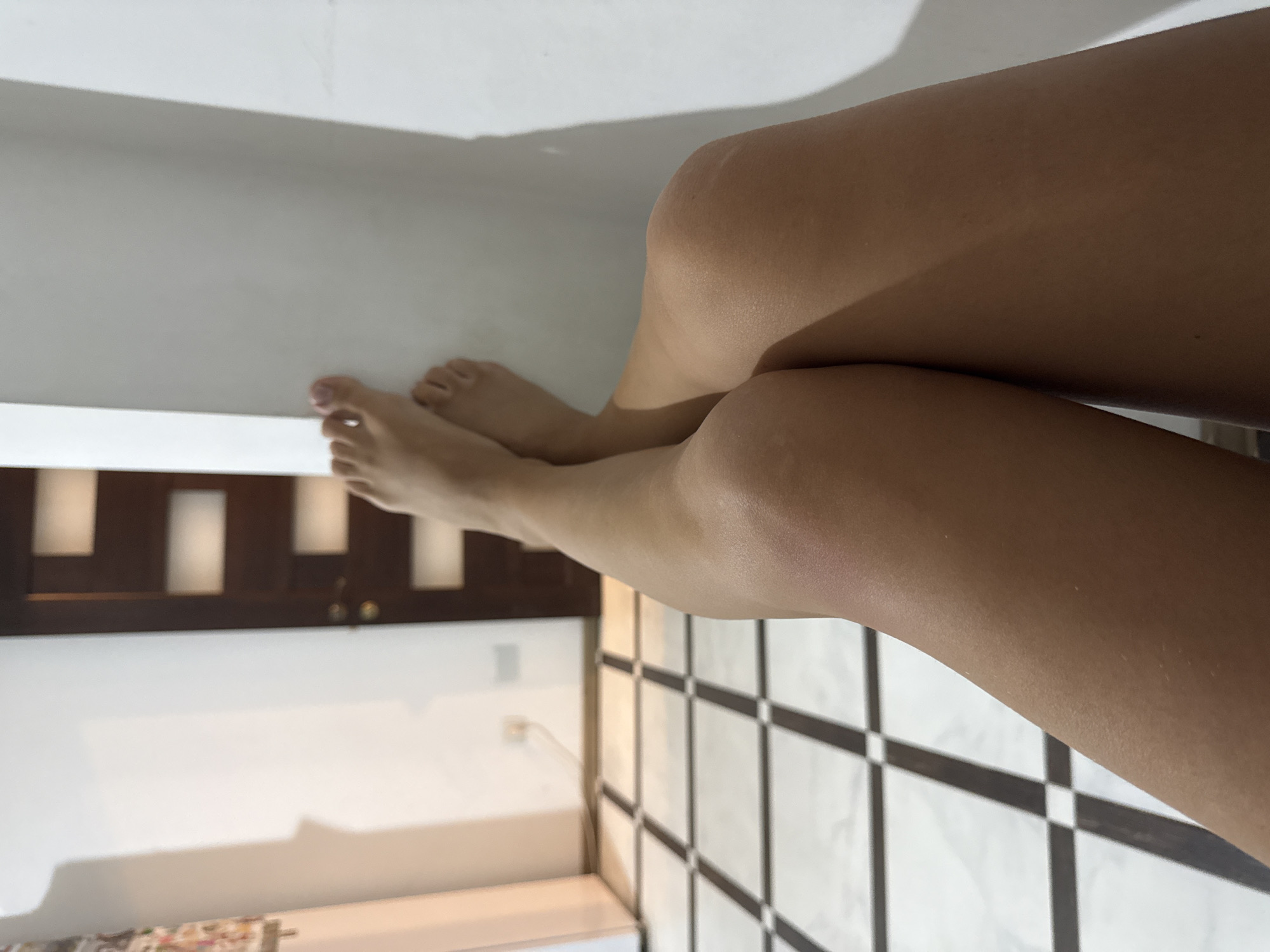 The image shows a person's legs, from the knees down to the feet, resting against a white wall. The legs are tanned and appear smooth. The feet are bare and positioned with toes pointing towards the left of the frame. The person's left leg is crossed over the right. The background consists of a tiled floor with a black and white checkered pattern, and a dark wooden door with several rectangular panels. A white wall forms the right boundary of the scene, with shadows cast by an unseen light source. There is a power outlet visible on the wall in the background. The image does not provide any specific details that would allow for identification of its location within Chișinău, Moldova.
