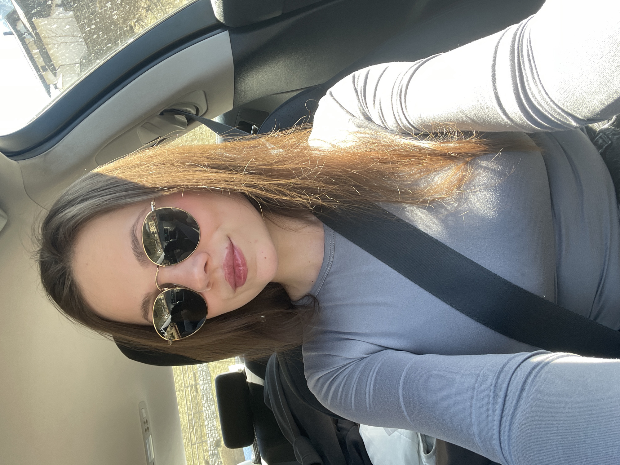 A woman with brown hair and wearing sunglasses and a gray long-sleeved shirt sits in a car, fastened with a seatbelt. The interior of the car and a glimpse of the outside environment through the car window are visible. The location is implied as Sappada / Plodn / Sapade, Italy.