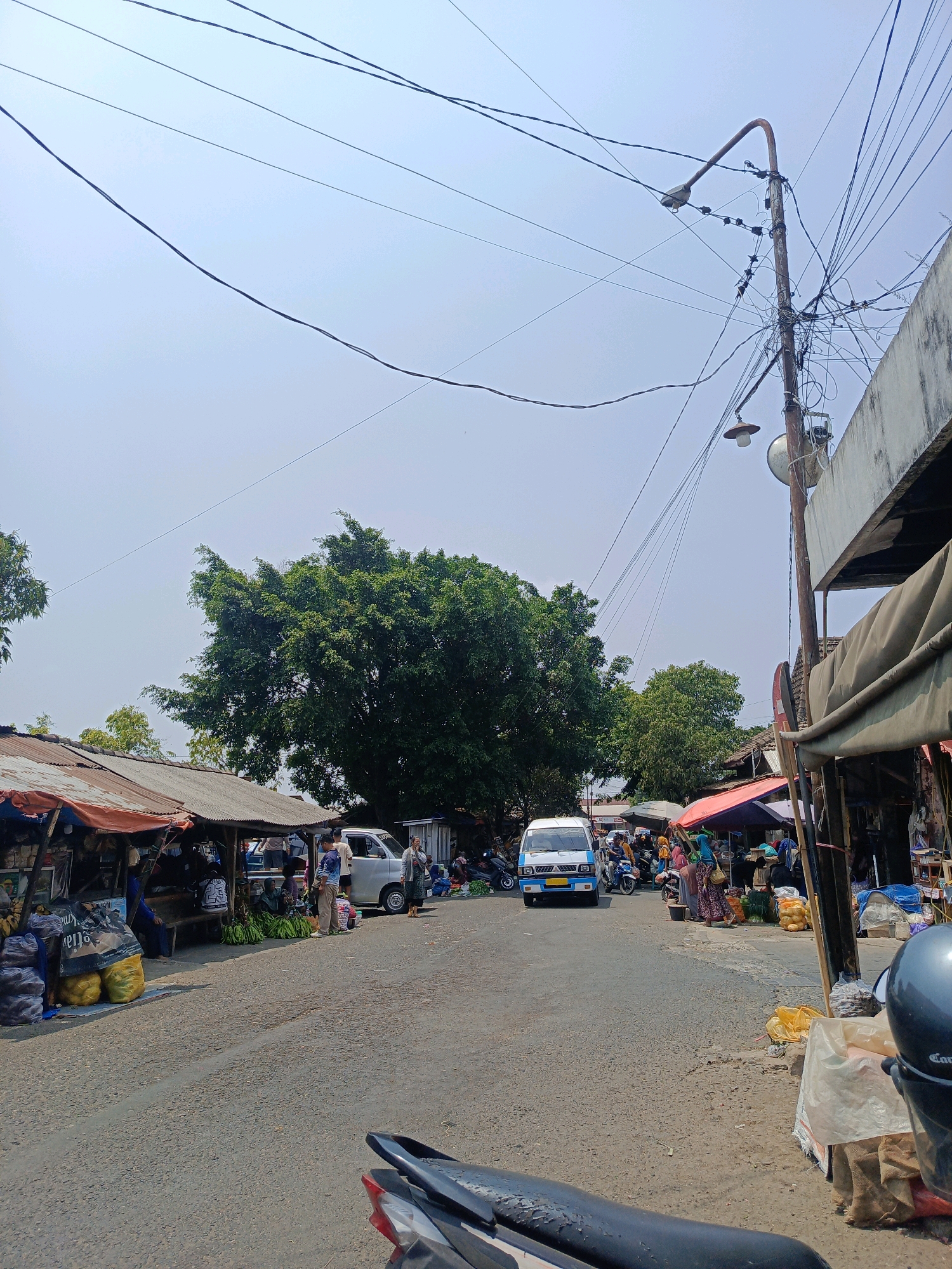 This is a photograph taken outdoors, likely on a street in the city of Sukowinangun, Indonesia, depicting a bustling marketplace scene. The sky is bright and appears hazy, characteristic of a sunny day in a tropical climate. A prominent utility pole with numerous electrical wires is situated on the right side of the frame, along with a streetlamp.
The foreground shows a portion of a motorcycle, suggesting the photographer is either on or near one. The road surface is asphalt, with visible tire marks and some debris.
Along the left side of the road, several market stalls are set up. These stalls have corrugated metal roofs and are laden with goods, including large sacks of produce (likely potatoes or onions, based on their yellow and grey colors), bananas, and other unidentifiable items. People are seen browsing or working at these stalls.
The center of the image features a paved road with vehicles. A white van is stopped in the middle of the road, and a smaller silver van is parked to its right. Several people are standing near these vehicles and the stalls. On the right side of the road, more market stalls are visible, also covered by tarps or awnings, displaying various goods including more sacks of produce and what appear to be food items.
In the background, a large, dense tree with green foliage stands out, providing a natural element amidst the urban market. Beyond the stalls and vehicles, more trees and some buildings are visible, indicating a populated area. The overall atmosphere is that of a lively, active local market.