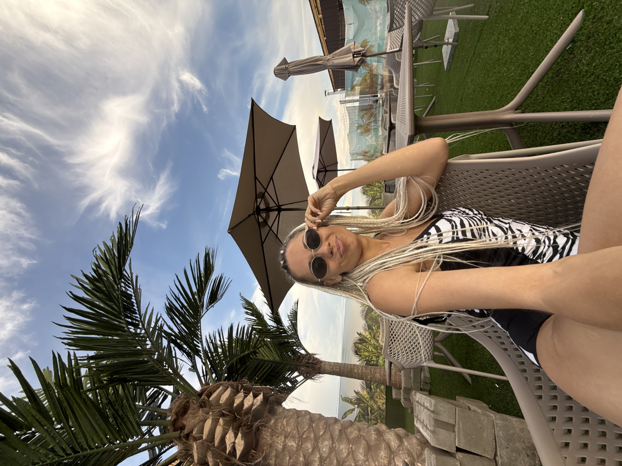 The image depicts a woman with blonde braids and sunglasses sitting on a chair in an outdoor seating area. She is wearing a black and white patterned swimsuit. Palm trees are visible in the background, along with umbrellas and tables. The ground appears to be covered in artificial turf. The sky is blue with wispy clouds. The setting suggests a tropical or resort-like atmosphere. There is no indication of the location being in Yaroslavl, Russia.