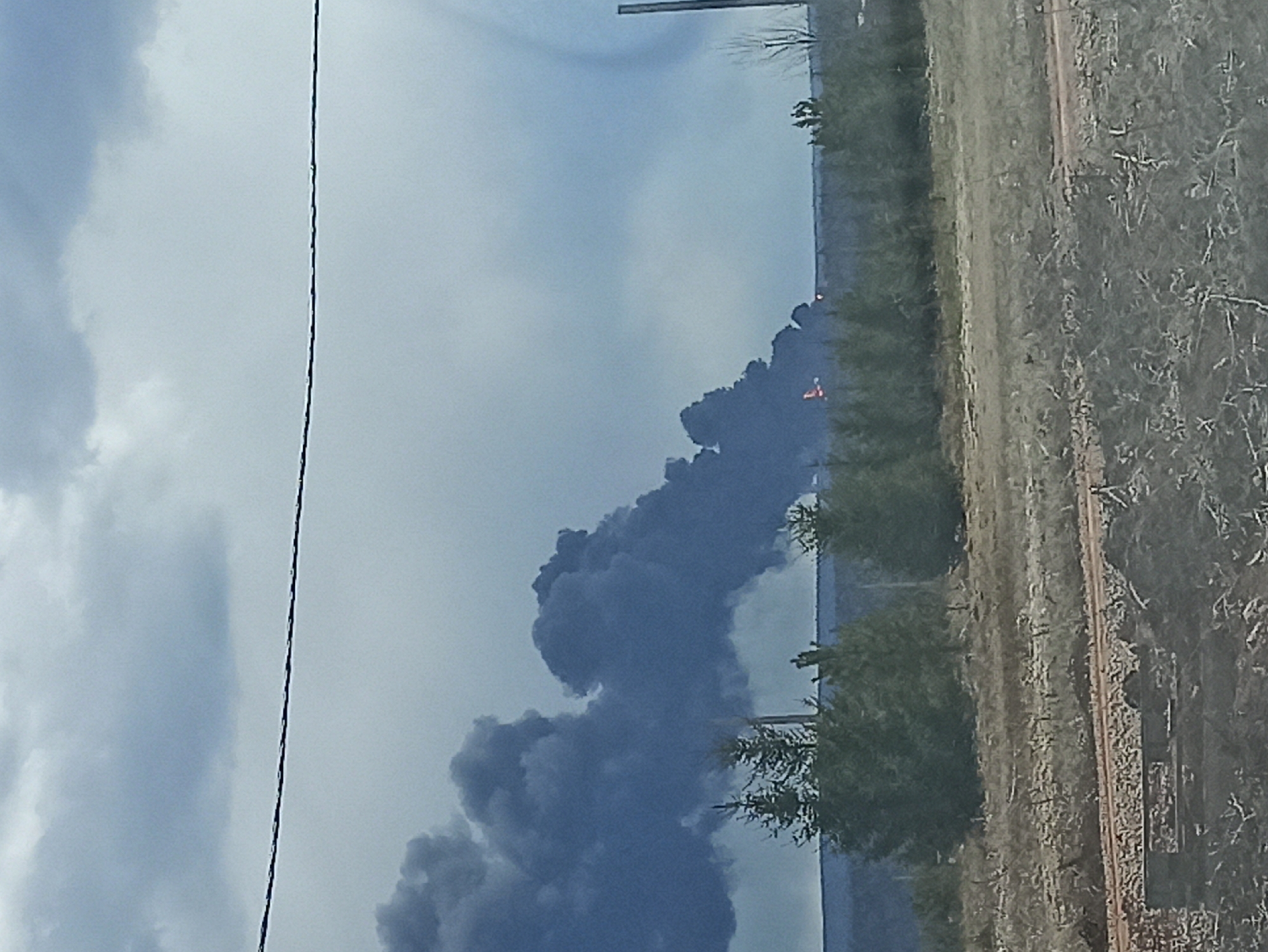The image displays a large, dense plume of dark smoke rising into an overcast sky, indicating a significant fire or industrial incident. The smoke occupies a large portion of the left and central frame, obscuring the background behind it.
The sky is cloudy with varying shades of light grey. A thin, dark, vertical line, possibly a utility wire, runs from the top to the bottom of the frame on the left side.
Along the horizon line, which is visible beneath the main smoke column, there is a strip of green and brown vegetation, appearing to be trees or bushes. Within the smoke-affected area, a small, bright orange or red object is discernible, its identity unclear but possibly a structure, equipment, or vehicle related to the smoke source.
The foreground and midground on the right side of the image show a dry, brushy landscape with a reddish-brown dirt path or road extending horizontally. The vegetation in this area consists of sparse, dry shrubs and branches.
The scene appears to be an outdoor, likely rural or industrial setting during daytime, characterized by the light sky. No specific location cues are visible, and there is no discernible text.