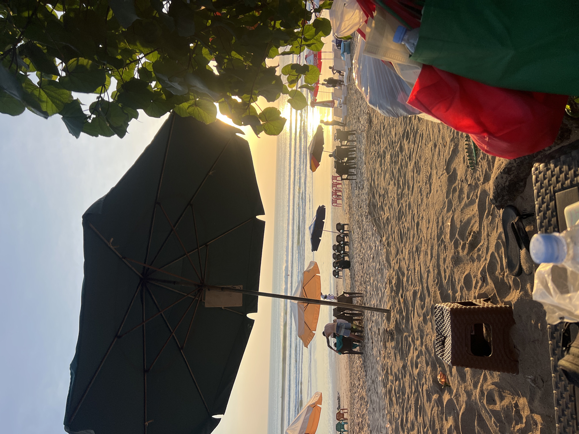 This is a medium shot of a beach in Kuta, Indonesia, during sunset. The foreground is dominated by a large, dark green beach umbrella and a cluster of colorful shopping bags, one red and one green, filled with items. A small, brown plastic stool is positioned in the sand to the left of the bags. In the midground, several beach umbrellas are set up, with some tables and chairs underneath. A few people are visible, including a man in blue shorts and a man in a dark shirt sitting at a table under an orange and white umbrella. Further in the background, the ocean stretches to the horizon, with gentle waves and a few surfers. The sun is setting, casting a warm, golden glow across the water and the beach, with lens flare visible through the leaves of a tree on the right side of the frame. The sky is a pale blue, transitioning to a soft yellow near the horizon. The sand is light brown with visible footprints and ripples.