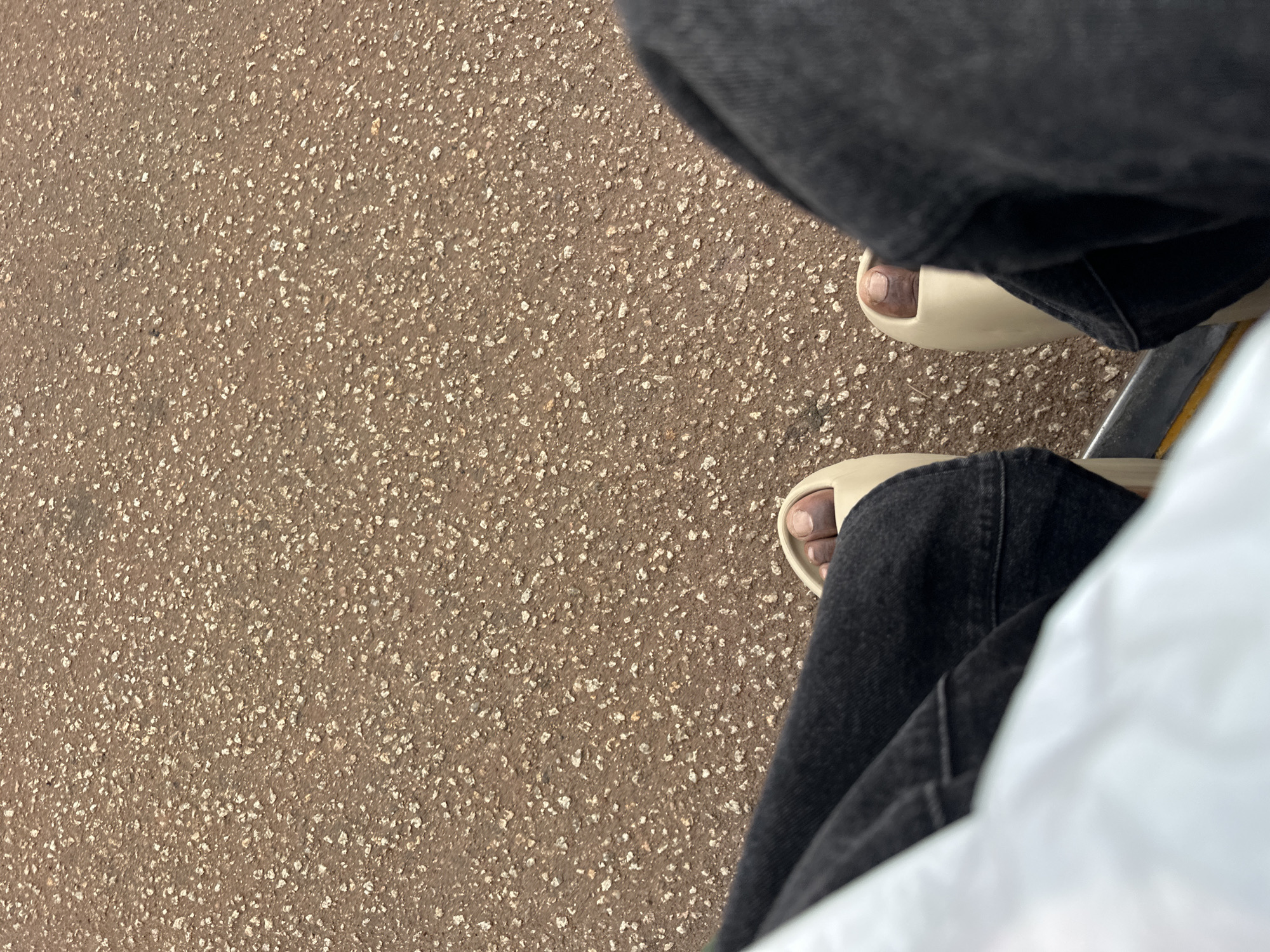 This media file shows a close-up view of a person's legs and feet, resting on a textured, gritty surface that appears to be asphalt or concrete. The person is wearing dark-colored jeans and light-colored slides. The background is predominantly the rough, speckled ground.
The perspective is from above, looking down at the individual's lower body. The lighting suggests it is daytime, as there are no strong shadows and the overall scene is well-lit. There are no specific cues to indicate the weather, but the outdoor setting is implied by the ground texture. No text is visible in the image. The scene is simple and focuses on the immediate surroundings of the person's feet and legs, offering no discernible emotional tone or specific event. The context provided places the location as Bukuru, Nigeria.