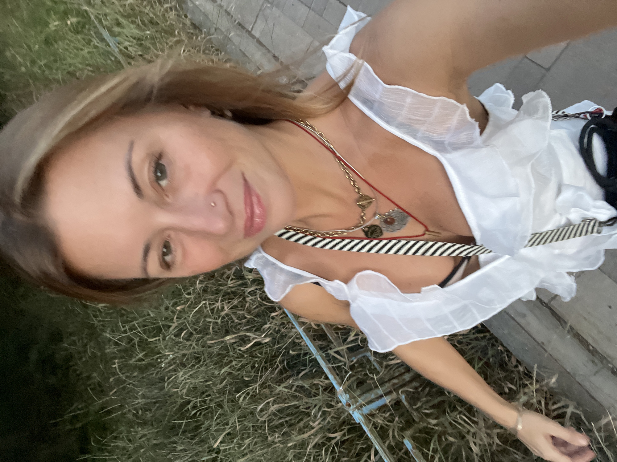 The image shows a medium shot of a woman with light brown hair, wearing a white ruffled dress and multiple necklaces. She is standing outdoors, with a blurry background of dry grass and a wooden walkway. A blue railing is visible to her left. The lighting suggests it is either dusk or nighttime. No specific details about the city of Samara, Russia are discernible from the image.