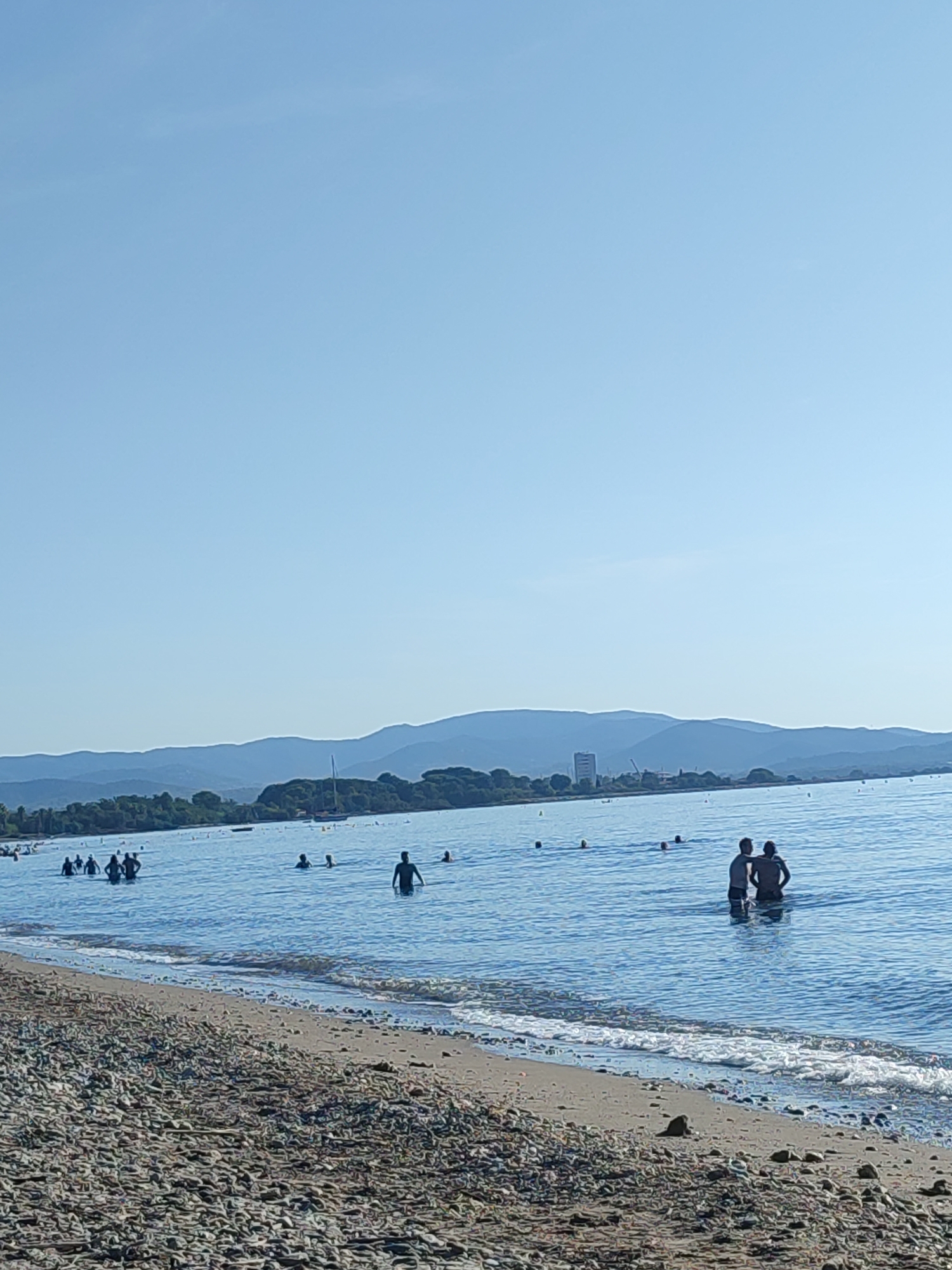 In Hyères, France, on a clear day with a pale blue sky, a wide, calm sea is visible. The foreground is a textured beach of small, multi-colored pebbles. Gentle waves lap at the shore. In the water, several people are wading and swimming. Two figures stand facing away from the camera, appearing to converse. In the middle ground, a scattered group of swimmers enjoys the water. A distant, tree-lined shore is backed by rolling, blue-toned hills and mountains. A sailboat and a few buoys dot the water. A modern, multi-story building stands on the shoreline amidst the greenery.