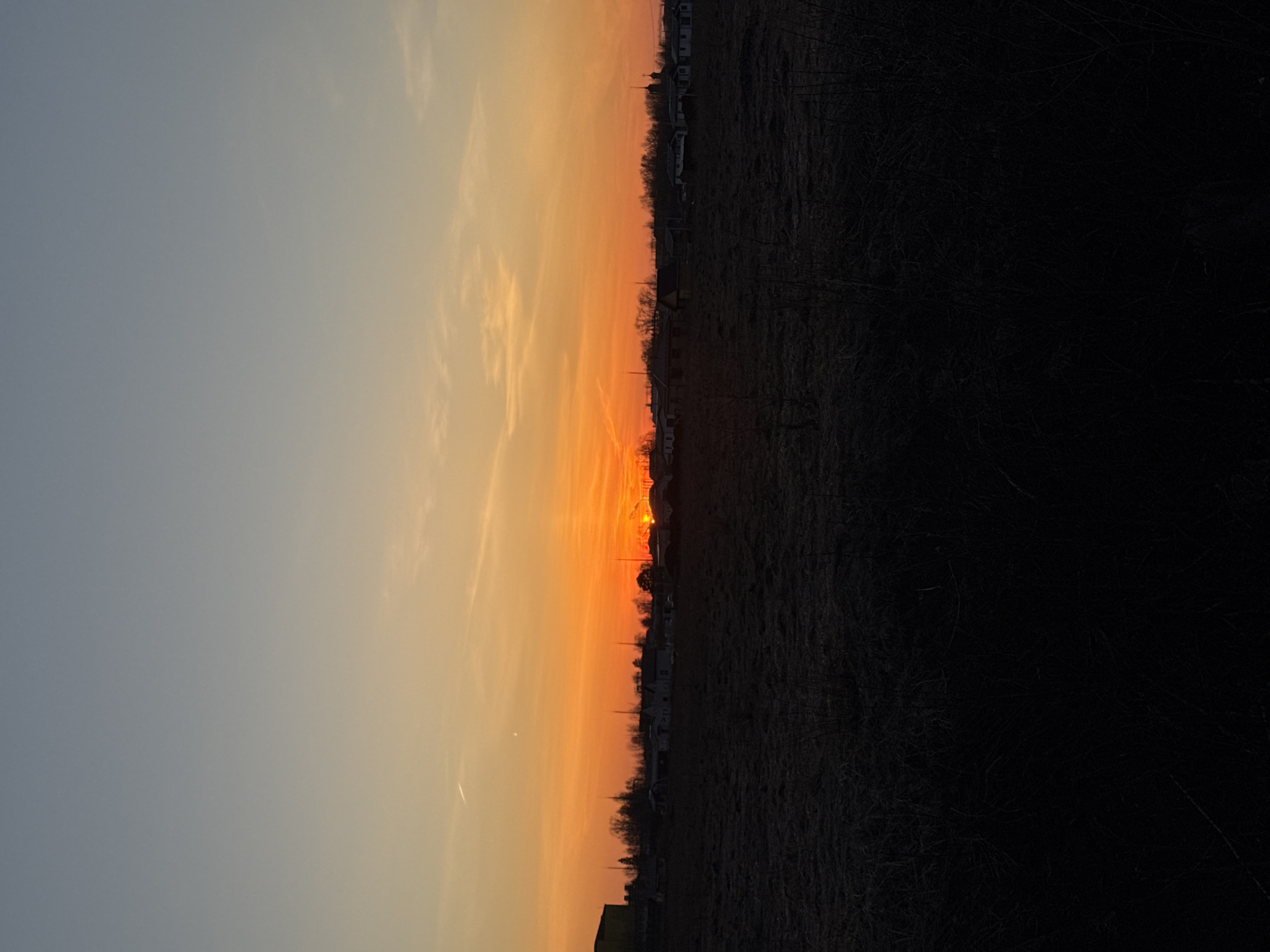 The image captures a sunset over a landscape in Горный, Russia. The sky transitions from a light blue at the top to orange and yellow near the horizon, where the sun is setting. The foreground is primarily dark, suggesting a field or unlit area. A row of houses or buildings is silhouetted along the horizon.