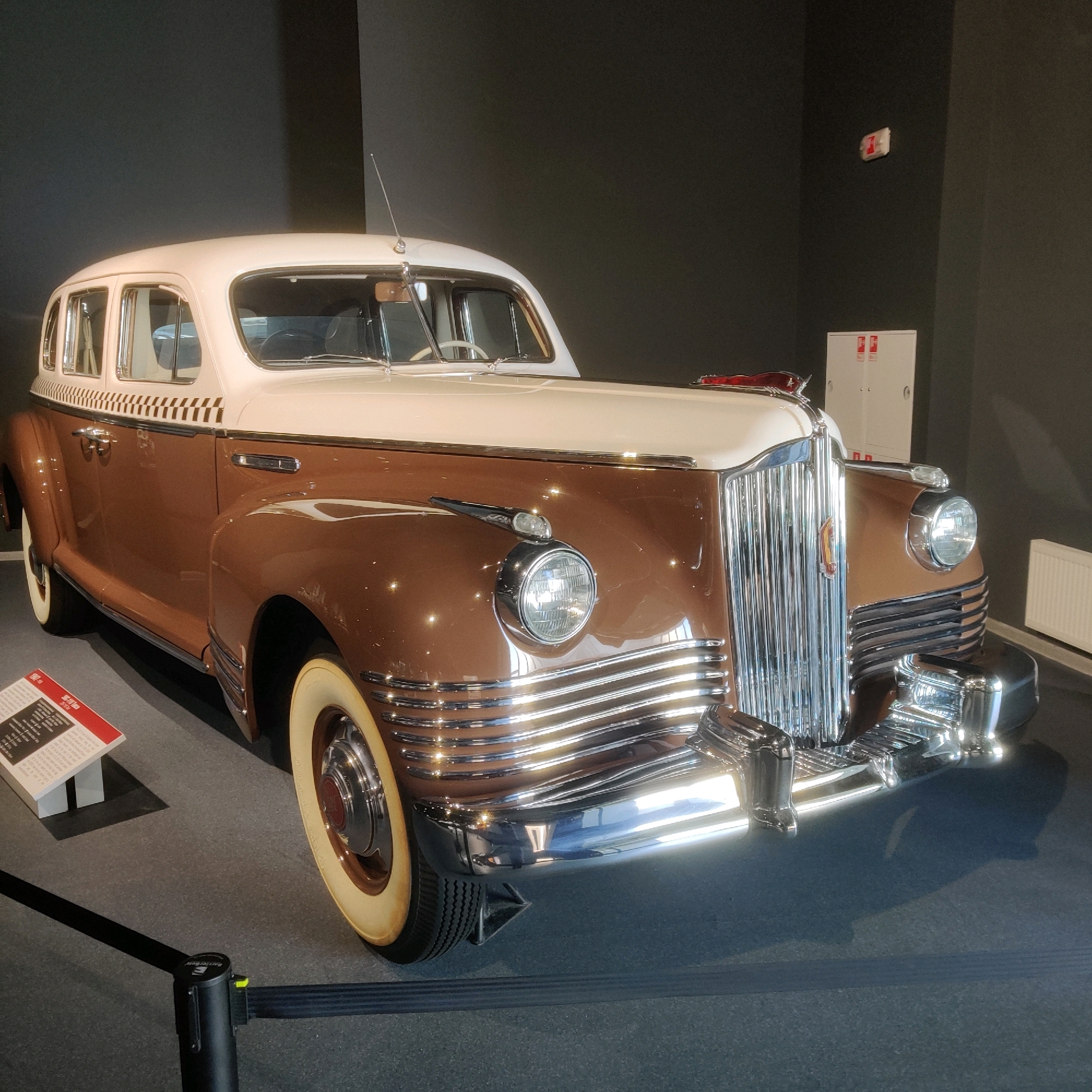 A vintage two-tone brown and cream-colored sedan is displayed indoors. The car has a chrome grille, rounded headlights, and whitewall tires. A small informational placard is placed to the left of the car. The vehicle is positioned on a dark gray carpeted floor, with a dark gray wall behind it. In the background, there are two white cabinets with red markings and a white radiator. The scene appears to be in a museum or exhibition space. Based on the informational placard which mentions "The History of Military Technology" and "Uralmash," this vehicle is likely located at the Museum of Military Technology UMMC in Verkhnyaya Pyshma, Russia.