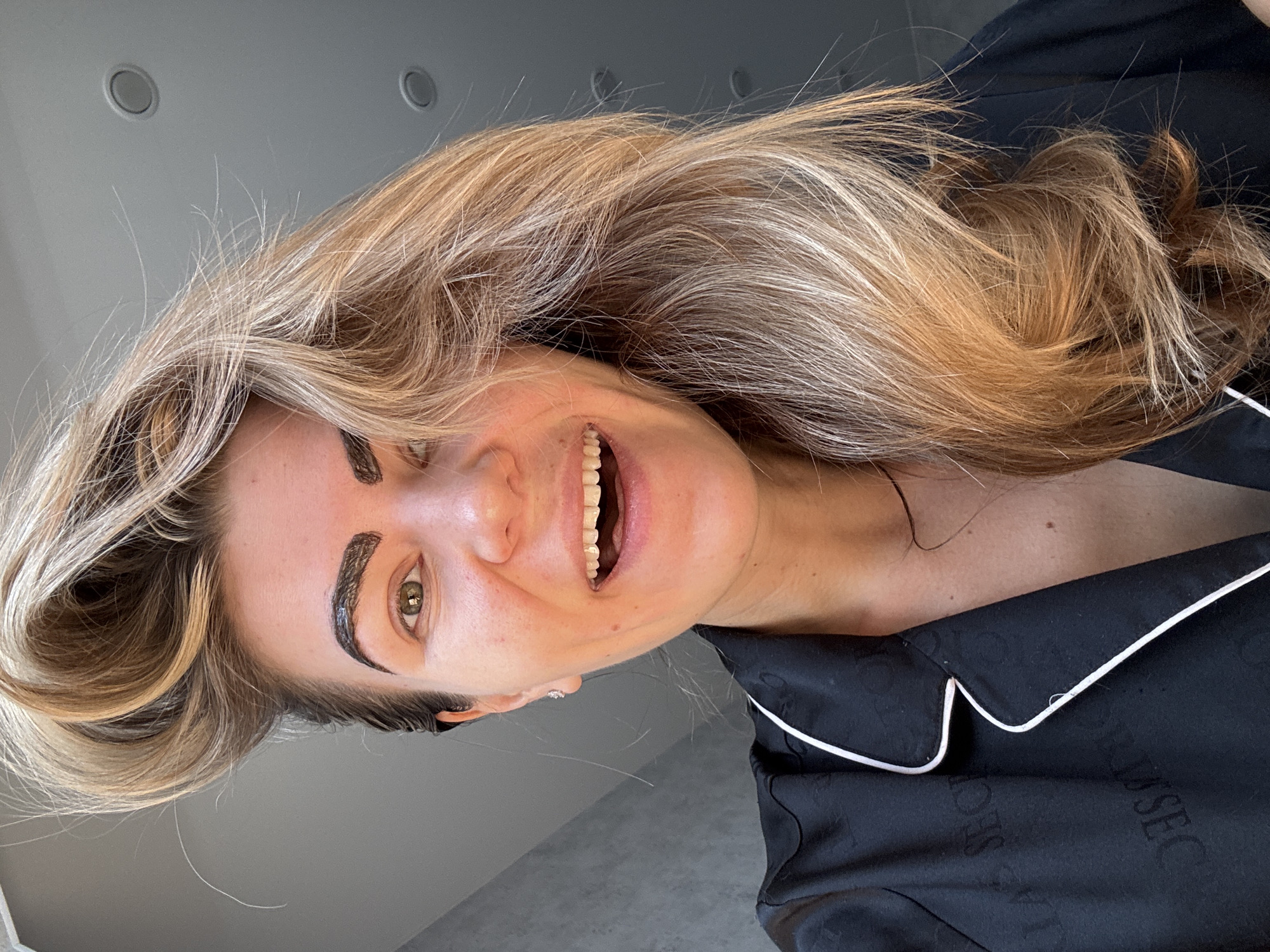 The image is a close-up of a woman with light brown hair, wearing a black pajama top with white piping. She is smiling and has dark treatment on her eyebrows. The background is a gray wall and ceiling with circular recessed lights. The location appears to be indoors, possibly a home, in an unspecified city and country.