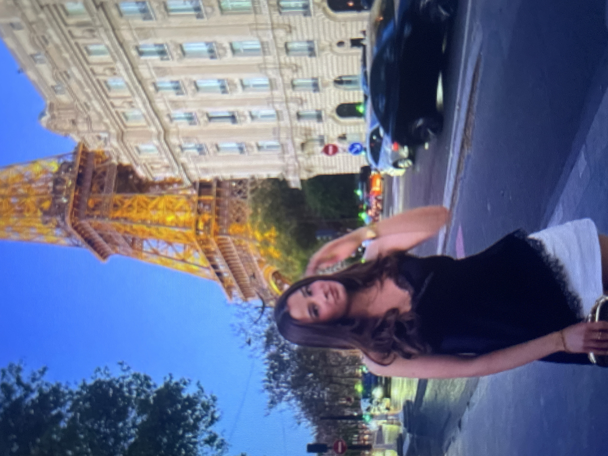 The image shows a young woman with long brown hair, wearing a black camisole with lace trim and white shorts, standing on a street with the Eiffel Tower in the background. She is holding a clutch purse. Cars and a building with ornate architecture are also visible. The sky is a deep blue, suggesting it is dusk or early evening. The image quality is low, with a noticeable pixelation. The location is identified as the city of Kołobrzeg, Poland, however, this is inconsistent with the presence of the Eiffel Tower, which is in Paris, France.