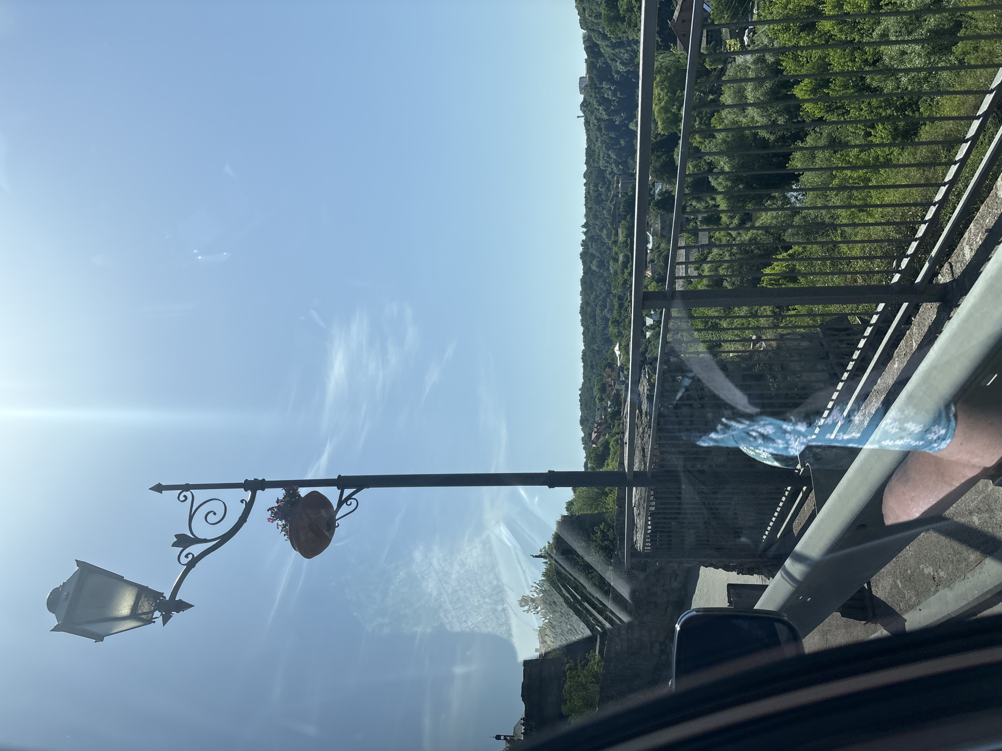 A street lamp with a decorative planter hangs over a stone wall. Beyond the wall, a lush green valley is dotted with houses and trees. The sky is a clear blue with a few wispy clouds. The reflection of the photographer and their car is visible in the glass of a window.