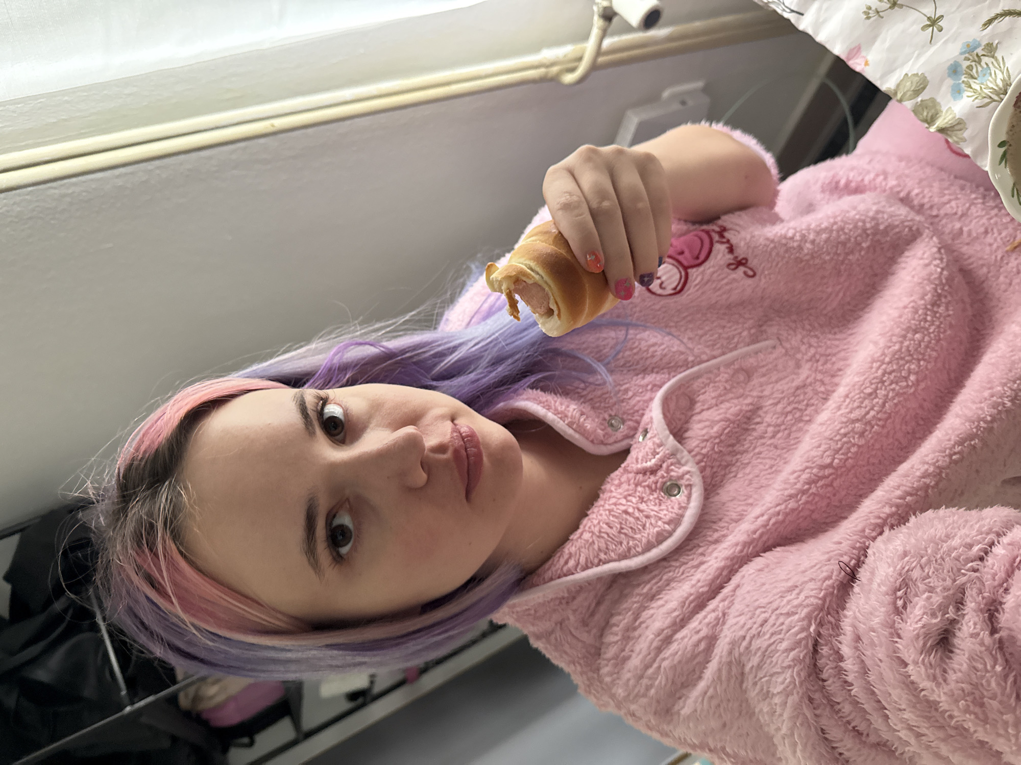 A female individual with light skin tone and bi-colored hair, dyed pink and purple with visible natural root growth, is depicted indoors, likely in a private residence within the City of Zagreb, Croatia. She is wearing a fuzzy, light pink long-sleeved top with white piping and snap buttons, featuring an embroidered design of two pink smiley faces and text on the left chest. Her right hand, displaying multi-colored nail polish, holds a partially consumed sausage roll. Her gaze is directed towards the camera with a neutral expression. The background to her left reveals an open shelving unit containing dark garments and a pink bag. To her right, a light-colored wall and a white curtain are visible. A floral-patterned bed sheet and a partial white bowl containing a dark substance are present in the lower right foreground.