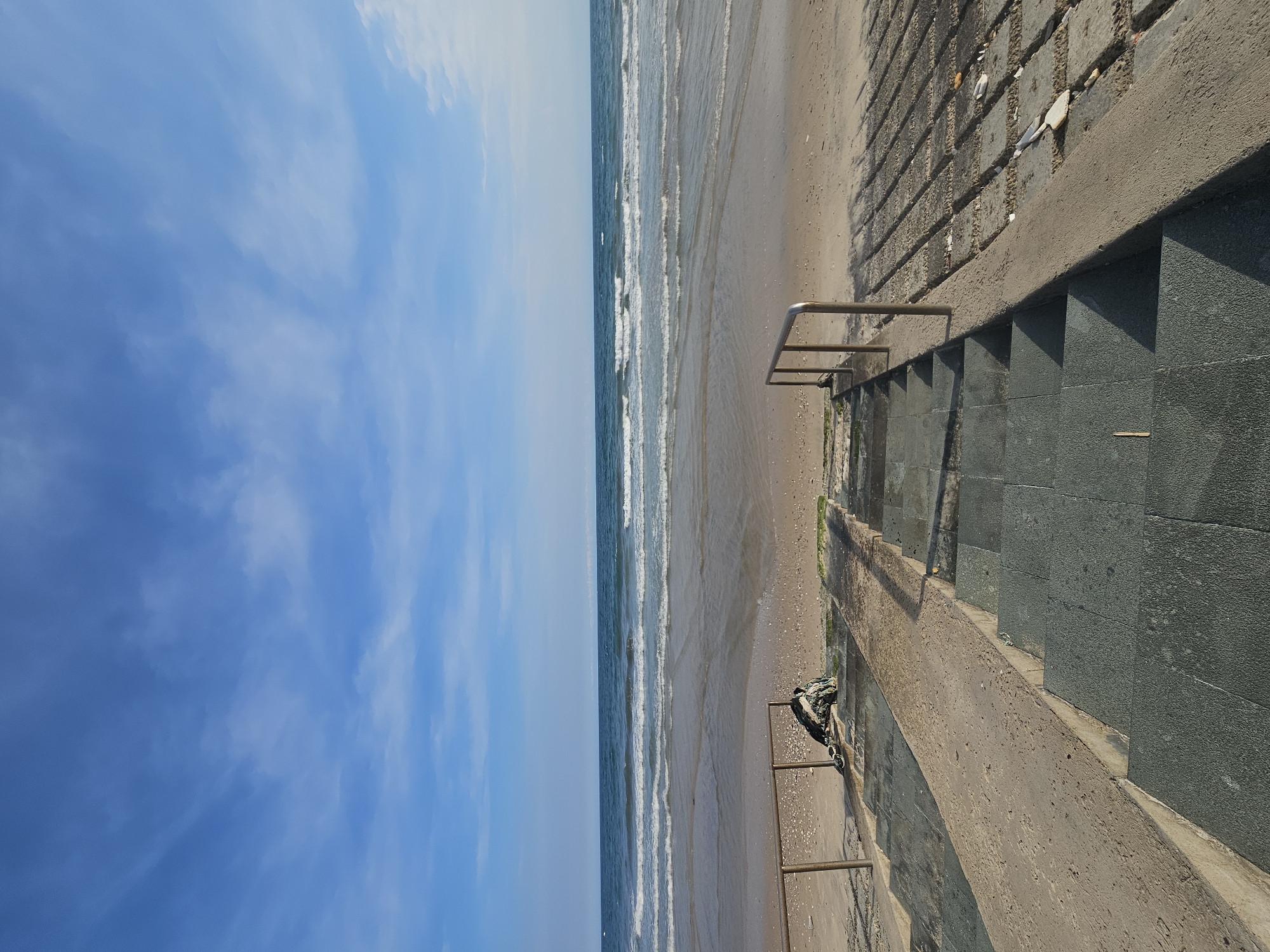 The image shows a view of the ocean from a concrete staircase in Mũi Né, Vietnam. The sky is blue with scattered white clouds. The ocean has moderate waves breaking on a sandy beach. The staircase is made of concrete and has metal railings. A pile of what appears to be fishing nets or fabric is on the left side of the stairs near the beach. The ground leading to the beach is paved with rectangular blocks. The overall scene depicts a typical coastal environment.