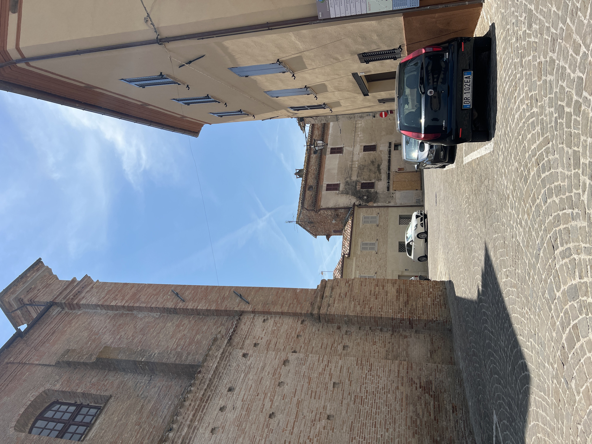 The image shows a narrow street in the town of Barbara, Italy, on a sunny day. The street is paved with cobblestones. On the left is a tall brick building with a large arched window. On the right is a lighter-colored building with blue shutters. Several cars are parked on the street, including a black Fiat Punto in the foreground. In the background, more buildings and a clear blue sky with some wispy clouds are visible. A street sign with Italian text is mounted on the building on the right.