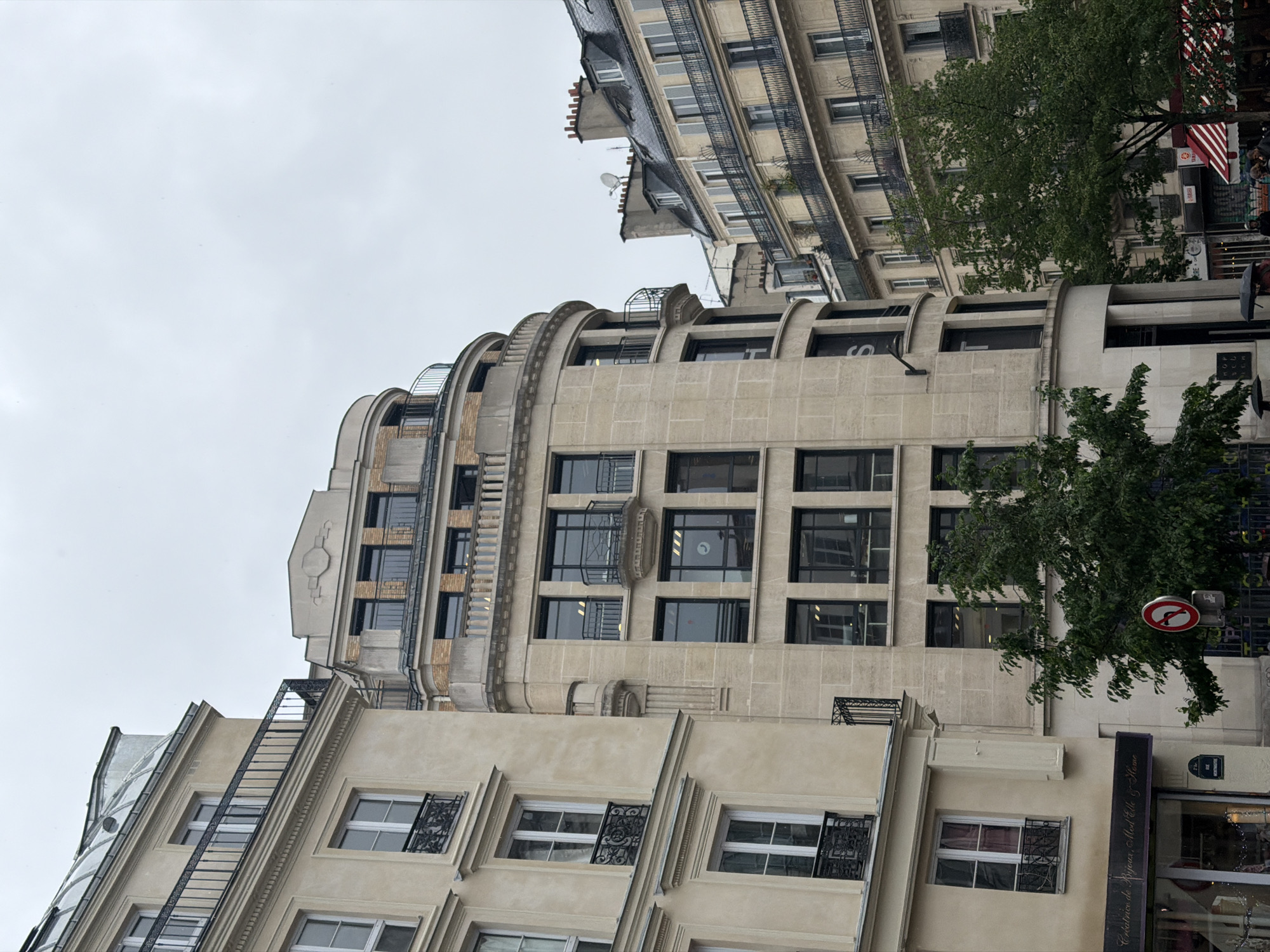 The image depicts a street scene in Paris, France, showcasing the architecture of the buildings. The focus is on a tall, light-colored building with a layered, curved design near the top. The building has multiple rows of windows, some with decorative ironwork. A fire escape runs along the side of the building. Trees are seen to the right, partially obscuring the building. There is a traffic sign attached to a pole near the tree. In the foreground, the bottom of a building with windows and a shop front are visible. The sky is overcast and grey.