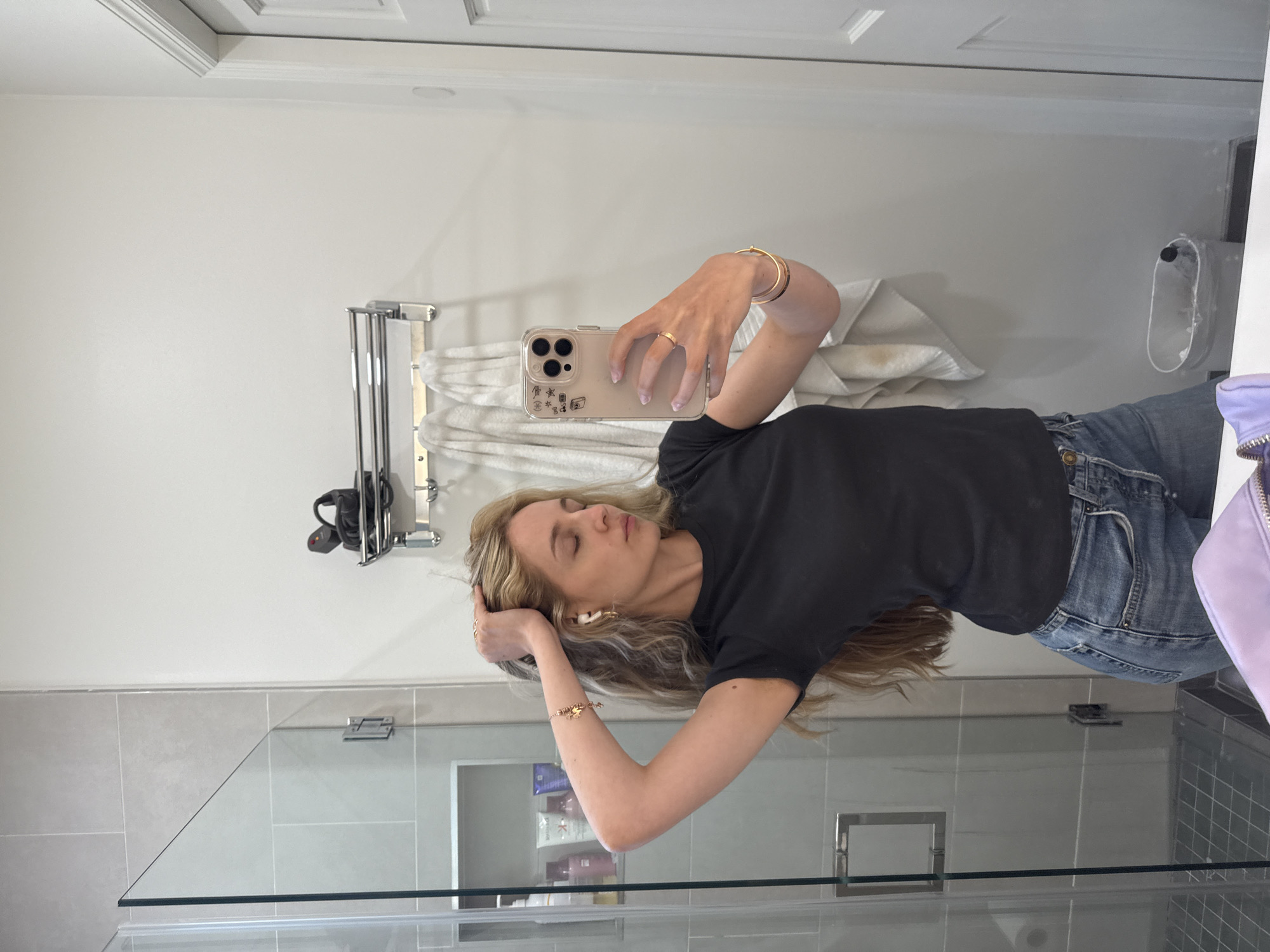 A woman with blonde hair is taking a mirror selfie in a bathroom. She is wearing a black t-shirt and blue jeans, and is accessorized with gold bracelets and earrings. She has her right arm raised and is touching her hair, with her eyes closed. The bathroom features a glass shower stall, tiled walls, and a towel rack with towels hanging on it. A trash can and a purple pouch are also visible in the frame.