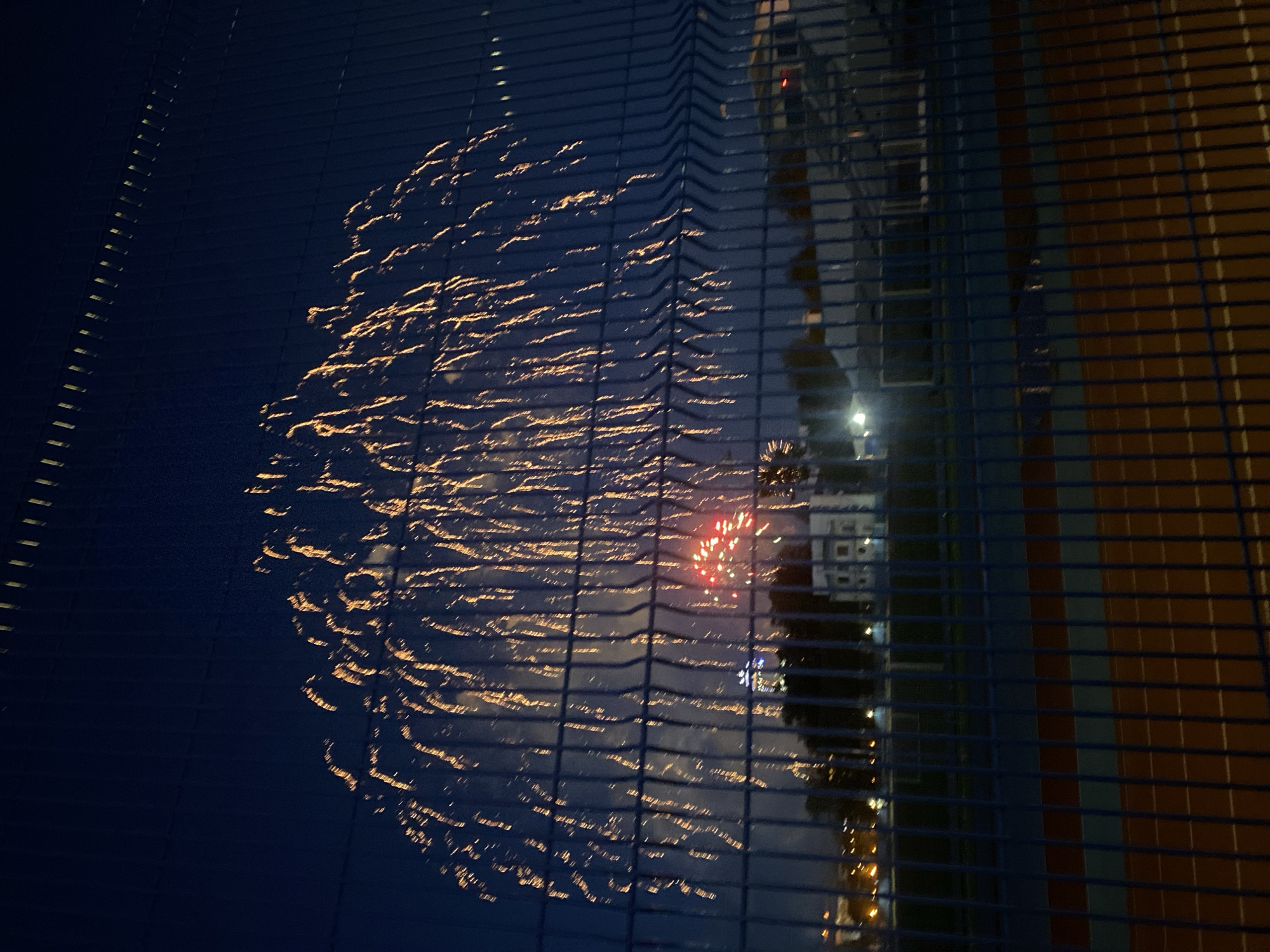 The image captures a firework display viewed through a metal fence. The fireworks are primarily gold and red, with trails illuminating the night sky. In the background, a building is visible. The location is likely Yekaterinburg, Russia, as indicated in the prompt, but without further information, it is impossible to determine the precise location within the city. The scene is dark, with the light of the fireworks creating a contrast against the night. No people are visible in the frame.