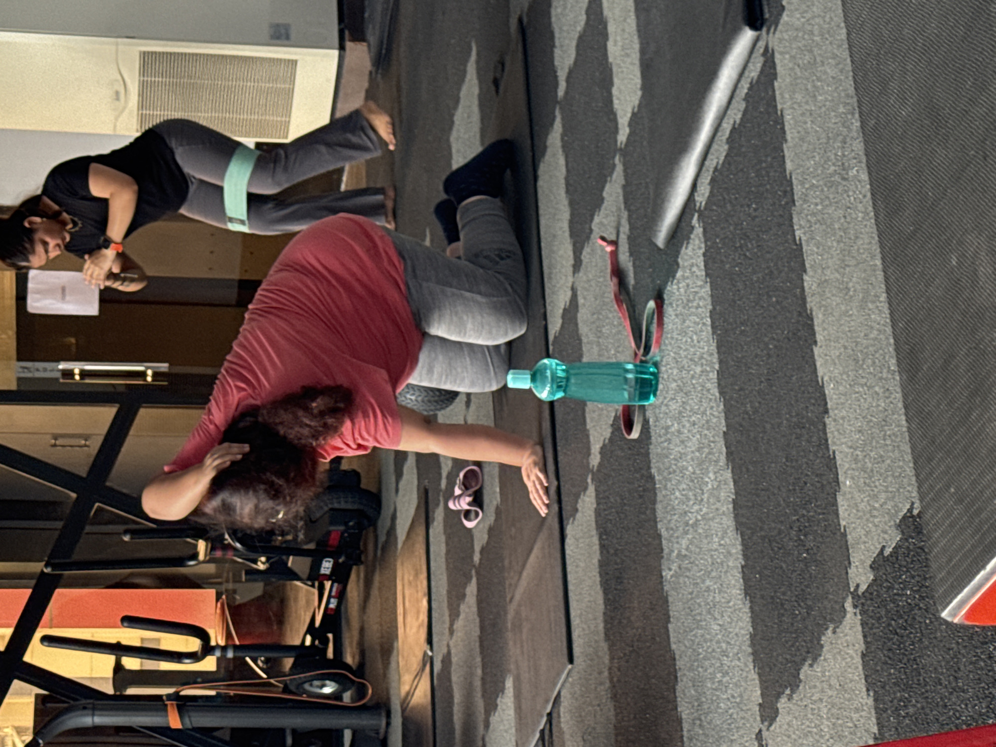 This image captures an indoor fitness setting, likely a gym or workout studio, featuring two women engaged in exercise. The floor is covered with distinctive interlocking grey and black patterned mats.
In the foreground, a woman wearing a pink t-shirt and grey track pants is positioned on her hands and knees, or possibly lying face down, with her head turned towards the left. Her dark, curly hair falls around her face, and her right hand is near her head. A teal water bottle and a pair of small pink sandals are visible on the mat near her. Behind her and to the left, gym equipment, possibly part of a rowing machine or exercise bike, is visible.
Further in the background, another woman stands, focused on her workout. She is dressed in a black t-shirt and grey track pants, with a light green resistance band positioned around her thighs, suggesting a glute or leg-strengthening exercise. She wears an orange fitness tracker on her left wrist. Her bare feet are visible. On the wall behind her, an air conditioning unit or vent is mounted.
The overall activity depicts individuals participating in a fitness routine. The lighting is artificial, characteristic of an indoor gym, with no indications of the time of day or outdoor weather. No discernible text is visible on any objects or surfaces within the frame. The location is provided as Kolkata, India.