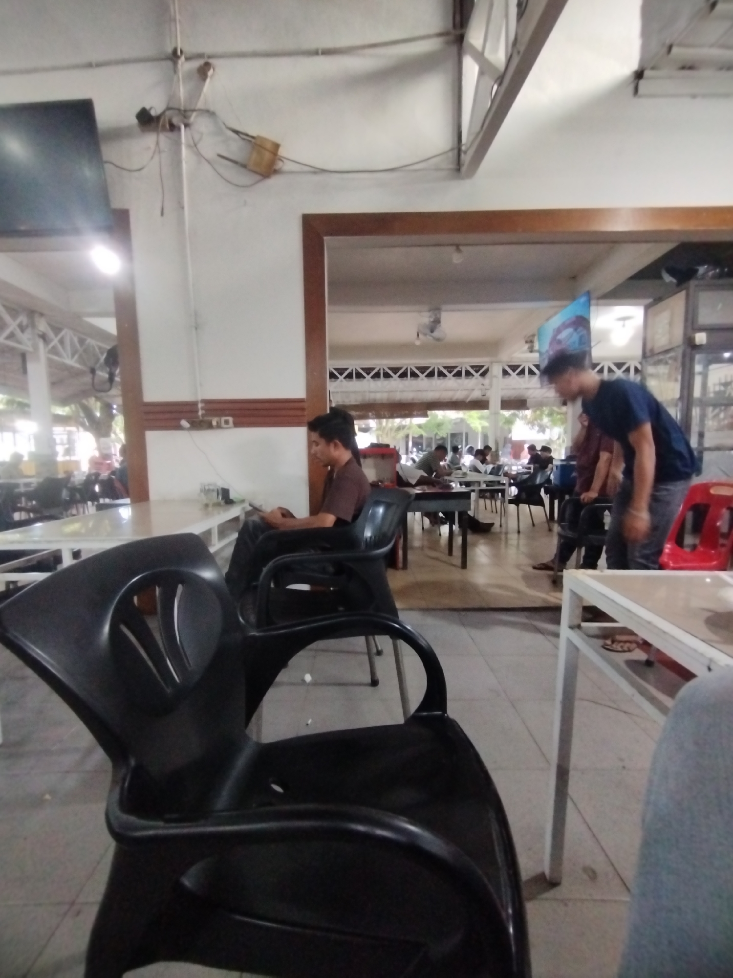 The photograph was taken in a café in Lhokseumawe, Indonesia. The scene features black plastic chairs in the foreground, one of which is prominently in focus. A man sits using a tablet in the middle ground near a large, white table. Other people are seated at tables in the background. A man stands in the right portion of the scene, partially obscured by a table and chair. The café has an open-air structure with high ceilings and white support beams. Wiring and a television are visible on the ceiling.