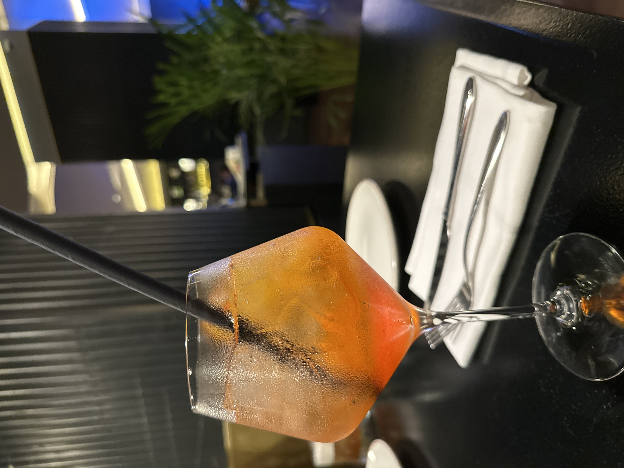 The image shows a cocktail glass filled with a reddish-orange liquid and ice, with a black straw inserted. The glass is set on a dark, possibly black, tabletop. To the right of the glass is a white napkin folded neatly, containing silverware (a fork and a knife). There are white plates visible in the background, and the leg of another glass is also present. In the background, some plants are visible, along with a blurry view of interior lighting, suggesting the setting is indoors, possibly a restaurant or bar. Based on the location details, this scene is likely in a restaurant or bar in Batumi, Georgia.
