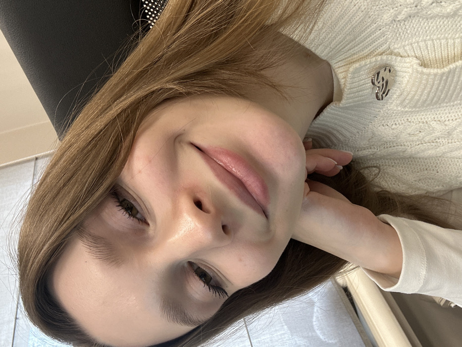 A fair-skinned woman with light brown hair smiles gently in a close-up selfie. Her lips are glossy, and her eyes are slightly squinted. She wears a cream-colored, textured sweater adorned with a silver brooch. Her hand lightly touches her neck. Part of a dark chair back is visible in the upper left corner.