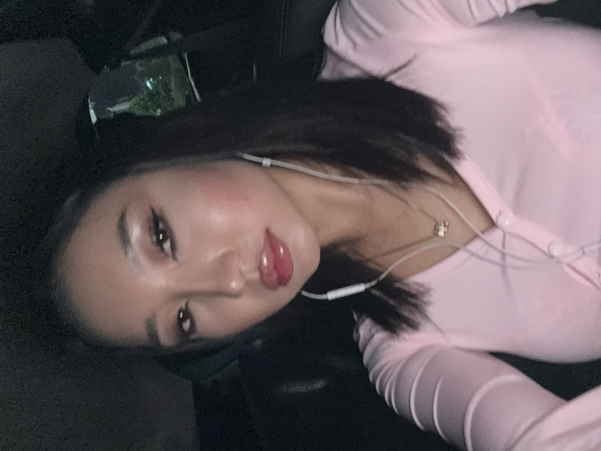 An overhead, close-up selfie of a young woman with dark hair and light skin is taken in a vehicle at night. She is wearing a pale pink, button-up, long-sleeved shirt and a delicate gold necklace. White earbuds are visible, with the wires draped over her shirt. Her makeup includes eyeshadow, eyeliner, and glossed lips. The background is dark, with a blurred reflection of trees and lights visible in the window behind her. The image does not contain any information that would allow for the determination of its location in Kuta, Indonesia.