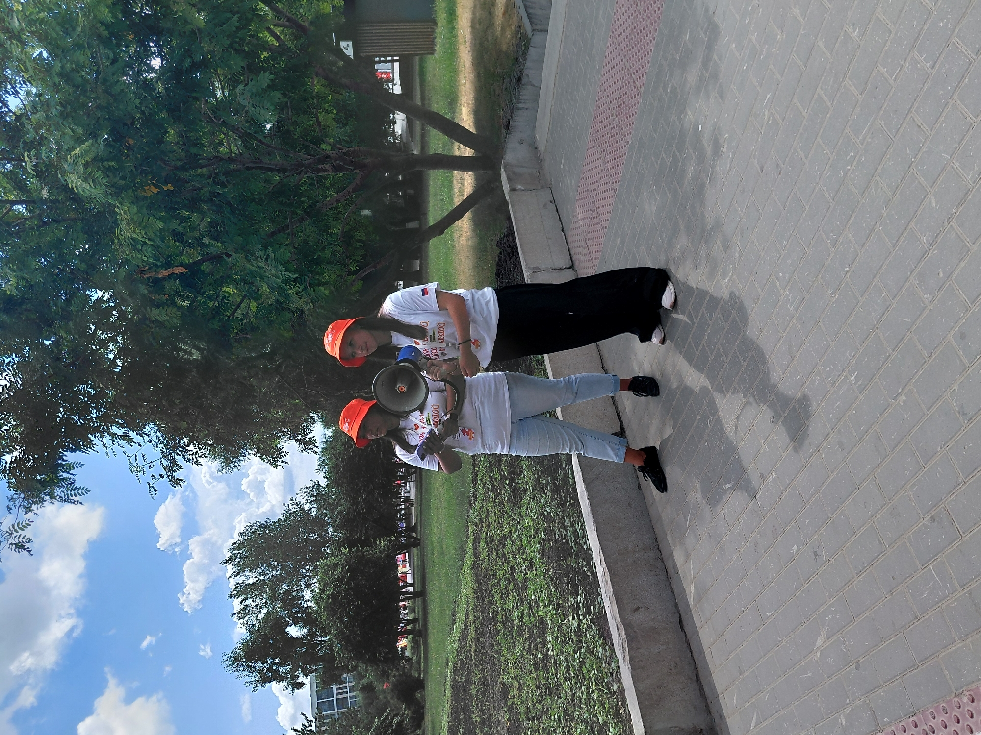 Two women stand on a paved walkway in a park-like setting in Novosibirsk, Russia. Both women are wearing orange bucket hats and white t-shirts. The woman on the left is wearing light blue jeans and black shoes, holding a megaphone in her right hand and a cell phone in her left. The woman on the right is wearing long black pants and white sneakers, also holding a megaphone. The background consists of grass, trees, and a cloudy blue sky. A concrete border separates the walkway from a grassy area.