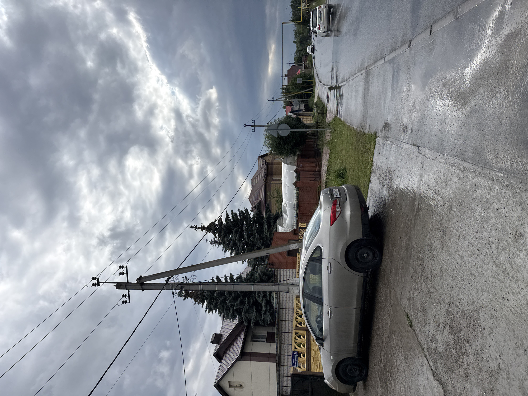 The image shows a street scene in Kazan, Russia, under a cloudy and overcast sky, suggesting it might be a dreary day. The ground is wet, indicating recent rain or ongoing drizzle, and puddles are visible on the asphalt road.
In the foreground, a light-colored sedan is parked on the side of the road, facing away from the viewer. The car appears to be clean despite the wet conditions. Beyond the car, the road curves to the right, with houses and fences lining one side. The houses have pitched roofs, and one visible house is light-colored with brown trim. A tall wooden utility pole with multiple wires runs along the side of the road, a common sight in residential areas.
Further down the road, two white cars are driving away from the viewer. The overall impression is of a quiet, residential street. The time of day appears to be daytime, given the visibility and the illumination from the sky, although the clouds obscure direct sunlight. The scene conveys a sense of dampness and muted light typical of a rainy or post-rain day. There is a visible street sign on a yellow post with Cyrillic text and a number, which is partially obscured but appears to read "Проспект Победы 37" (Victory Avenue 37).