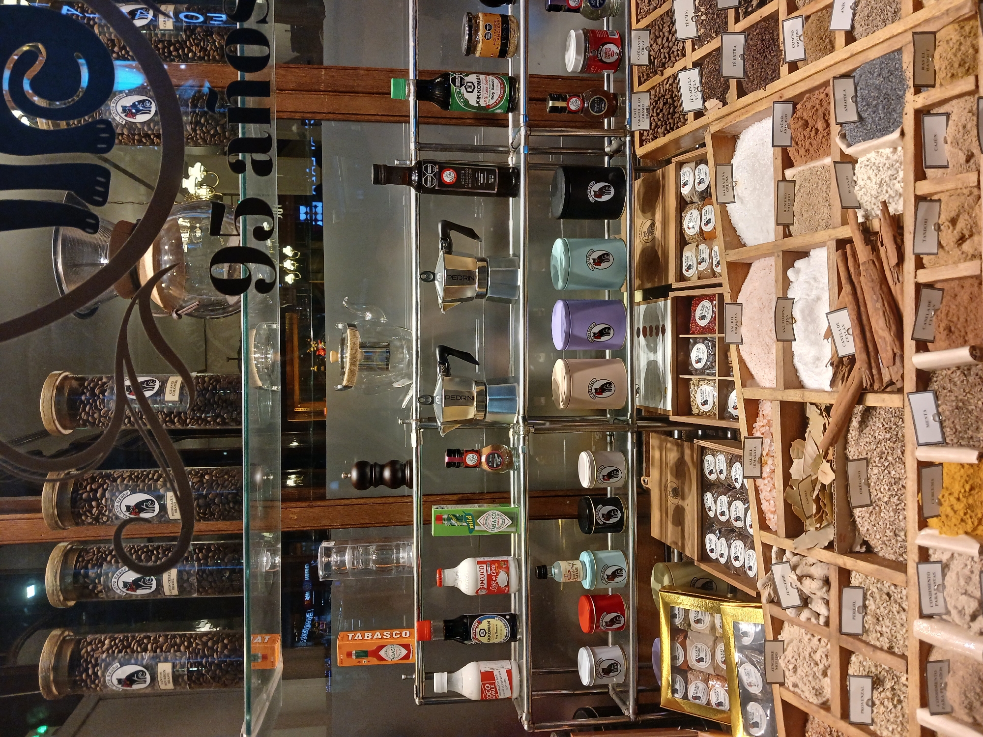 The image depicts the interior of a shop in Buenos Aires, Argentina, likely specializing in coffee and spices. In the foreground, various jars of coffee beans are visible, branded with "El Gato Negro." A glass display case contains bottles of vinegar, olive oil, soy sauce (Kikkoman), Tabasco sauce, and various coffee-making equipment like Moka pots. To the right, there is a wooden shelf unit filled with assorted spices, each with a label. The number "95" is prominently displayed on the glass window, potentially signifying an anniversary of the shop "El Gato Negro."