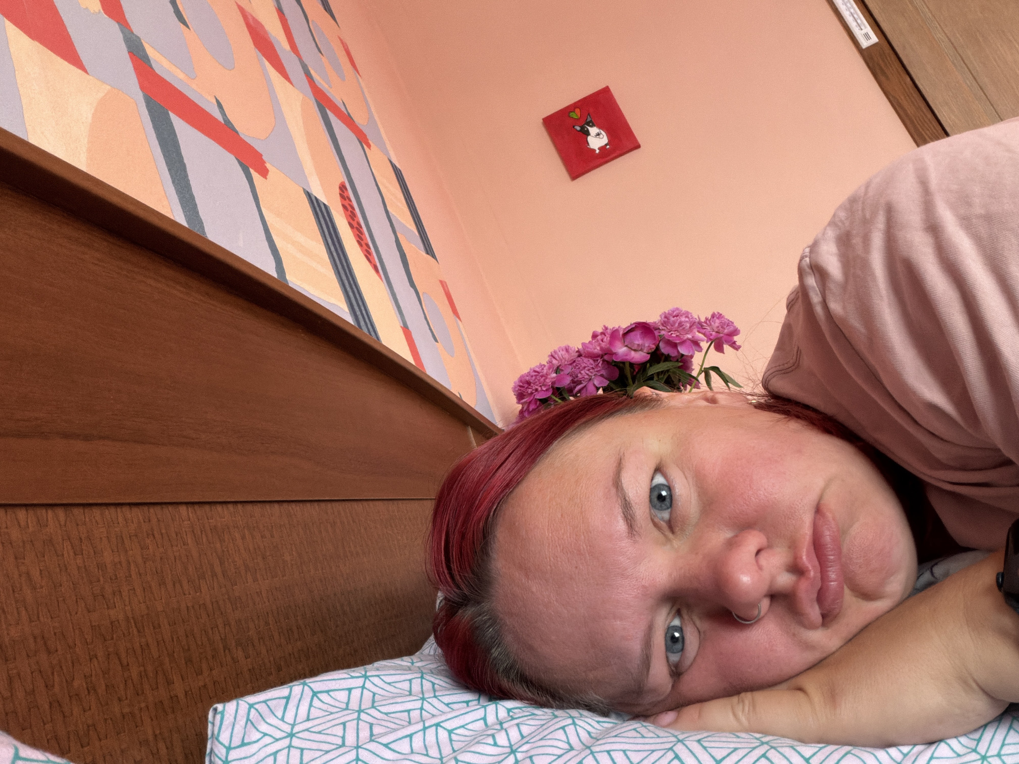 A close-up shot depicts a person lying on a bed with a blue and white patterned pillowcase. The person has red hair and light blue eyes, with a nose ring visible. The person's head is adorned with pink flowers. The background includes a light pink wall with a small painting of a dog. To the left is a wooden headboard and a wall with an abstract orange, red, gray and beige design. The location may be in Chaikovsky, Russia, but this cannot be determined from the image alone.