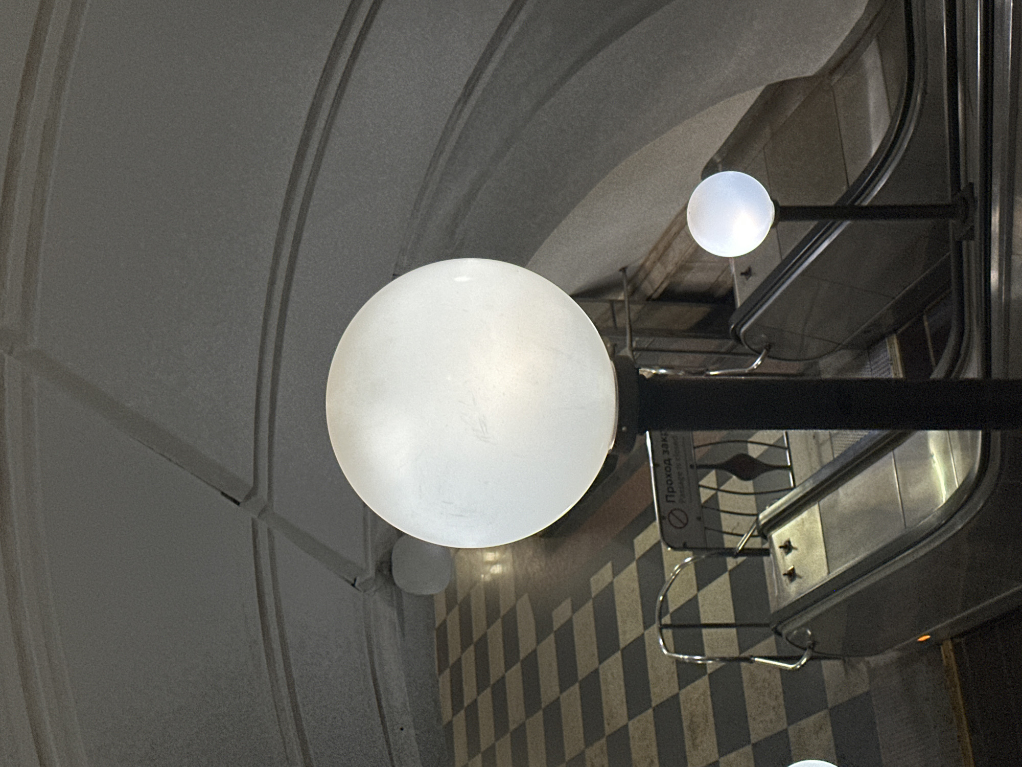 The image depicts an interior scene, likely in a subway station in Moscow, Russia, as suggested by the architectural style and details.
The primary focus is on two spherical light fixtures mounted on a black metal frame. The larger one is in the foreground, while a smaller one is visible in the background.
The background consists of a tiled floor with a checkered pattern. An escalator is visible to the right of the frame, suggesting the location is a transit hub. Signage with text in Russian can also be observed near the escalator.
The ceiling is arched and features a panelled design, contributing to the impression of being in a subway station.