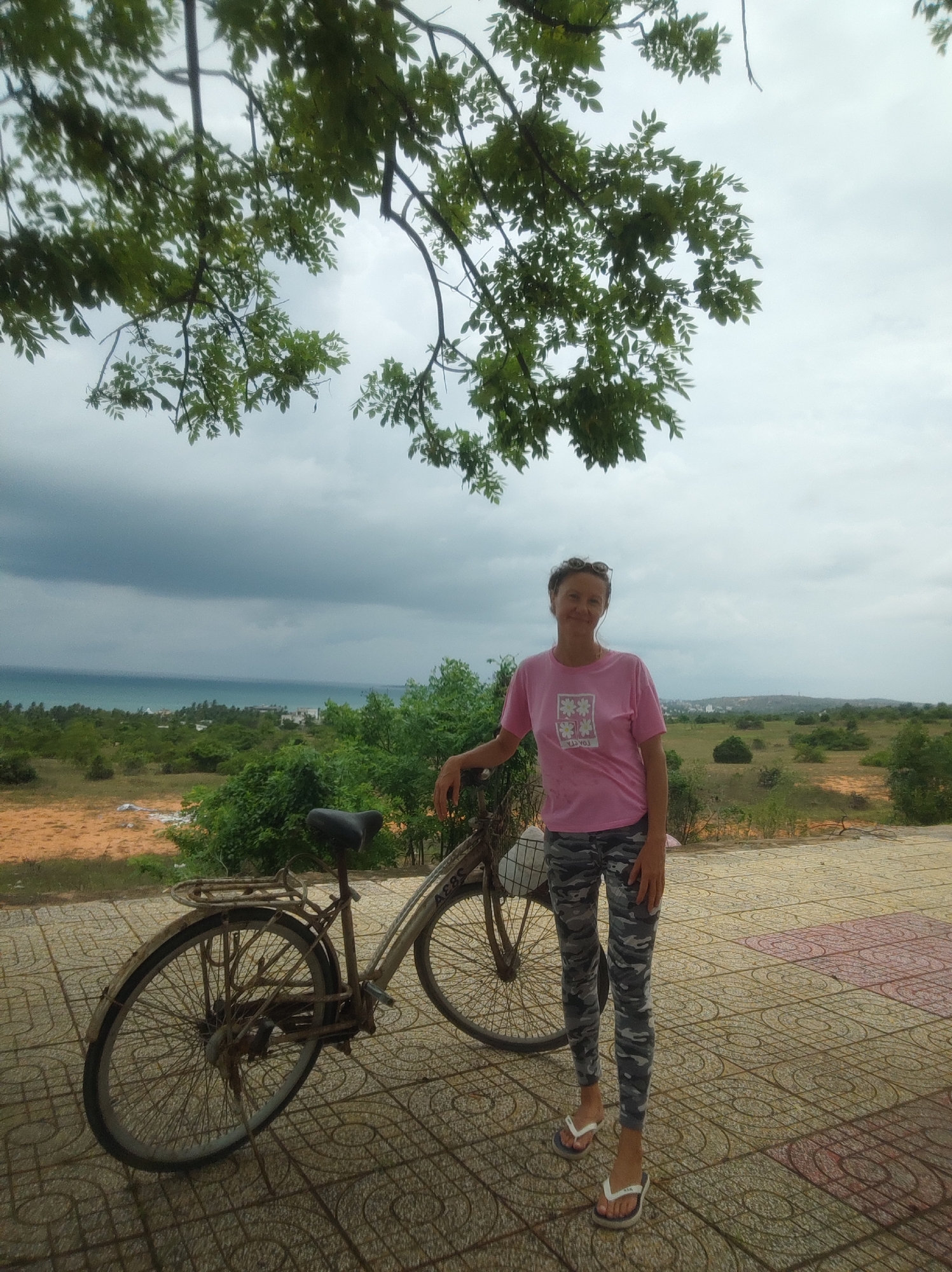 An outdoor, full-shot photograph captures a woman standing beside an old bicycle, set against a panoramic coastal landscape under an overcast sky. The woman, appearing middle-aged, wears a pink t-shirt adorned with four white floral squares and mirrored text that reads "YOUTOG" (or similar letters). She also wears camouflage-patterned leggings and white flip-flops, with sunglasses resting on her head. She offers a slight smile to the camera, with her right hand gently resting on the bicycle's handlebars.
The bicycle is an older, somewhat rusted model, equipped with a rear rack and a black seat. The letters "ABS" are visible on its chain guard.
The foreground is a paved area featuring patterned tiles in shades of terracotta and greyish-brown. Behind the woman and bicycle, the landscape consists of dry, reddish-brown earth interspersed with sparse green bushes and trees, typical of a scrubland or semi-arid coastal region. In the mid-distance, a large body of water, likely the ocean, stretches under dark, heavy clouds that dominate the sky, suggesting an overcast day or an approaching storm. A distant coastline with faint structures can be seen across the water. The upper portion of the frame is partially obscured by the green leaves and branches of an overhead tree. The scene conveys a moment of repose or a scenic stop.