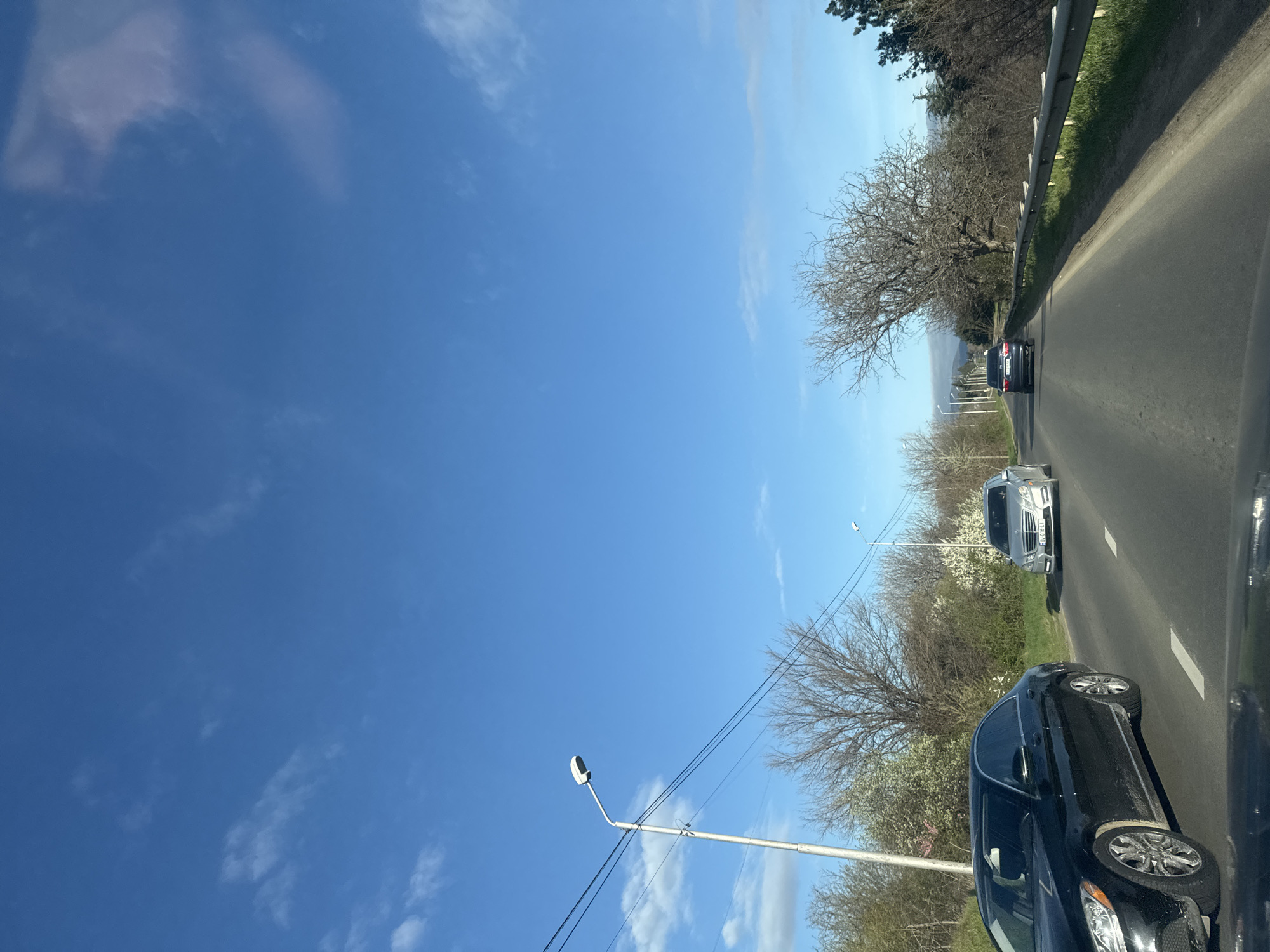 The image shows a road in Tbilisi, Georgia, with several cars driving on it. There is a black car in the foreground, followed by a white car, and a dark-colored car further down the road. The road is lined with trees and streetlights. The sky is blue with some clouds. There is a metal guardrail along the right side of the road.
