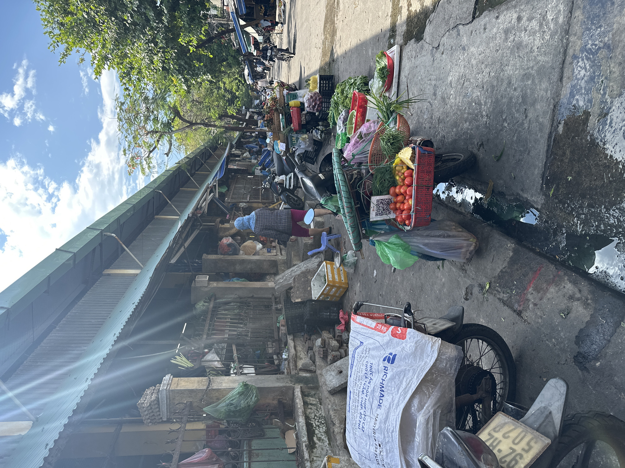 The image shows a street market in Đà Nẵng, Vietnam. Stalls with corrugated metal roofs line the left side of the street. People and parked motorbikes are visible. Vegetables and fruits are displayed for sale, some arranged on motorbikes. The street surface is concrete, with damp or wet patches. A large white sack with the word "RICHMADE" printed on it is visible on a motorbike at the bottom of the image. A vehicle license plate with the number "74-76" is visible. Trees and the sky with clouds are visible at the top of the image.