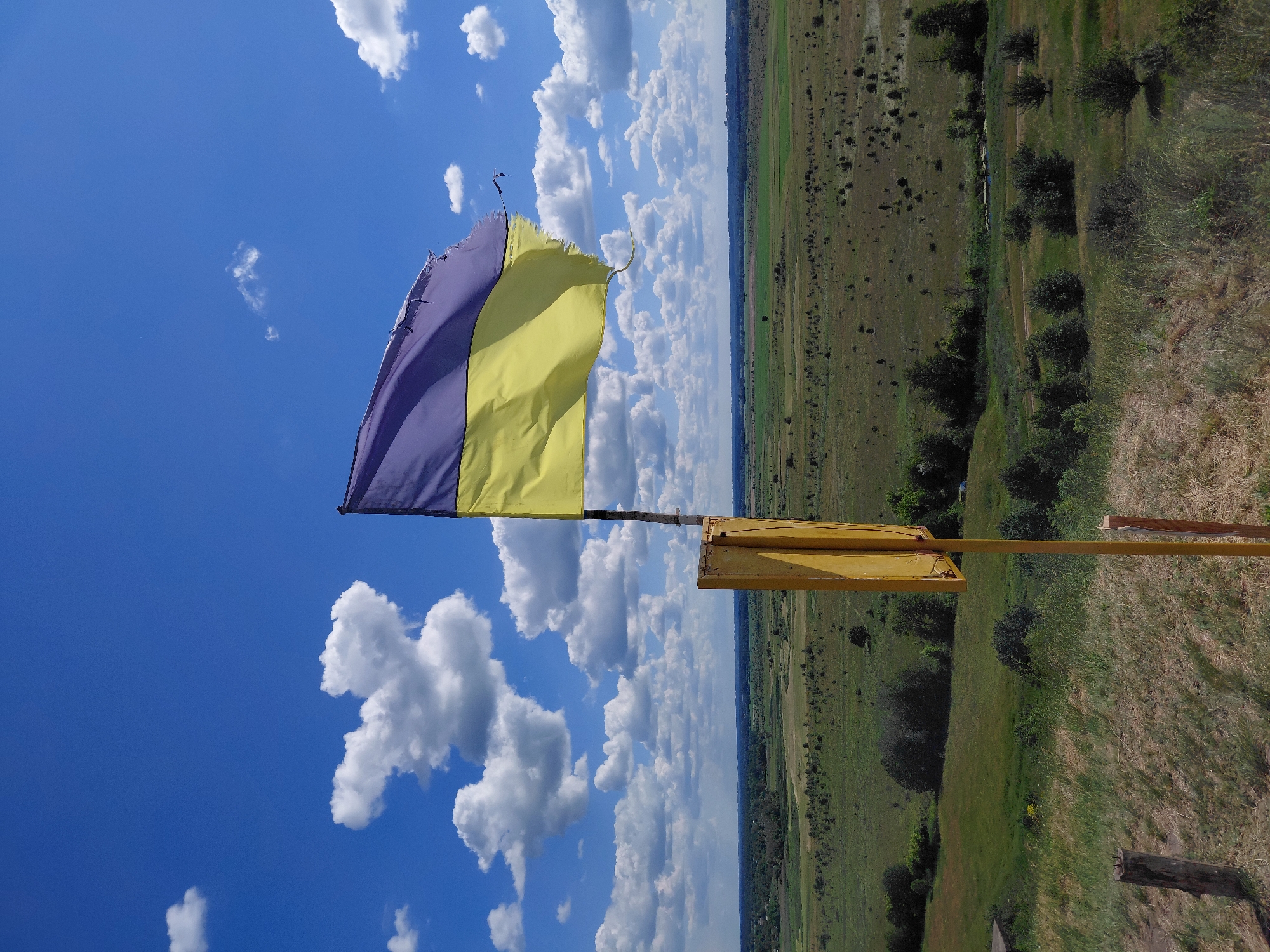 A slightly tattered Ukrainian flag flies on a pole in the foreground. The flag is divided horizontally into two equal parts, with blue on top and yellow on the bottom. The pole is a weathered yellow metal post with a square yellow sign attached to it. The flag and pole are positioned on a grassy, uneven hill. Behind the hill, a vast, green, rural landscape stretches to the horizon under a bright blue sky dotted with fluffy white clouds. The sky takes up roughly the top third of the image, and the landscape the bottom two-thirds.