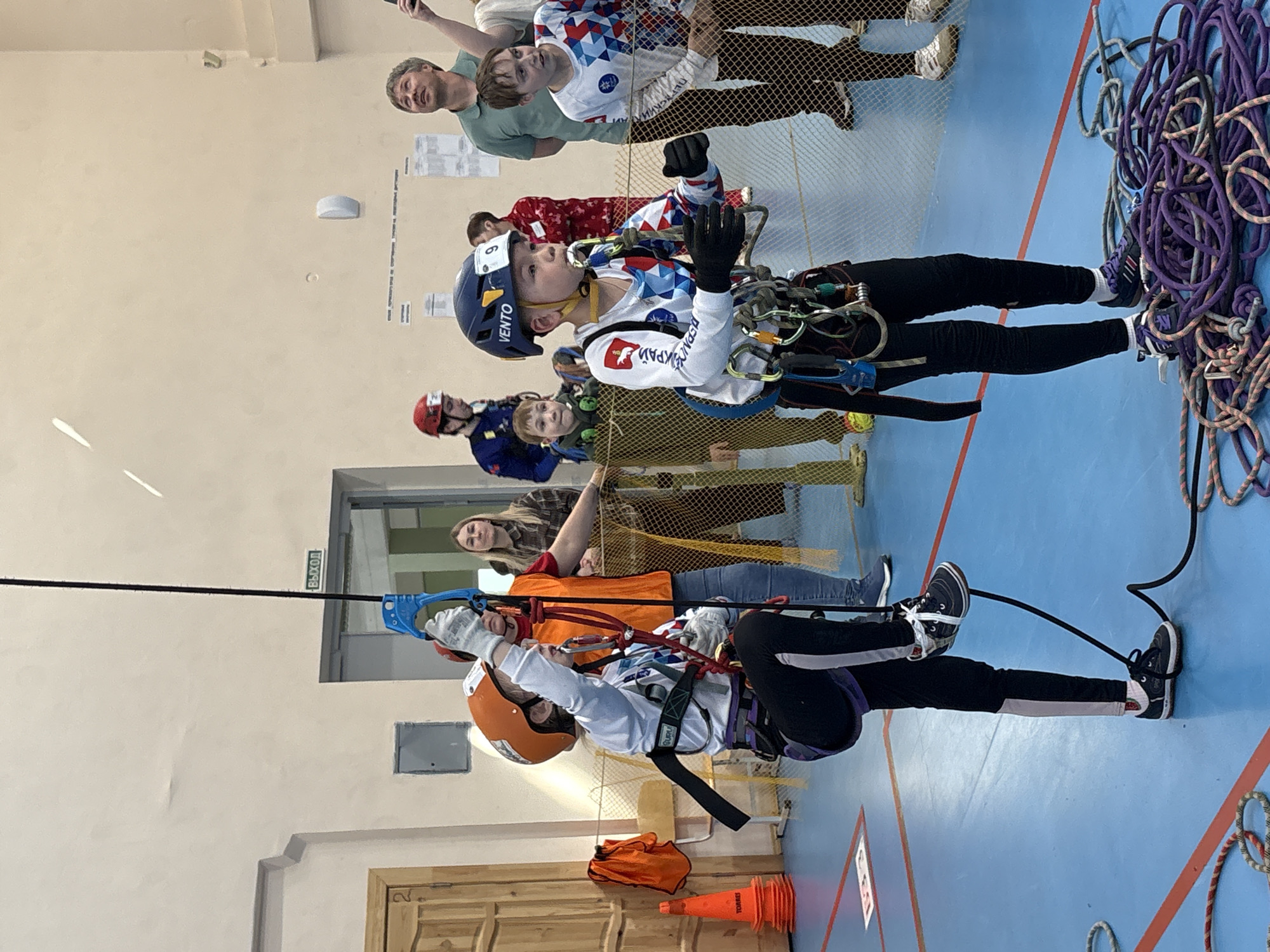 The image shows two children participating in a climbing exercise indoors. The location appears to be an indoor gymnasium or sports hall. The floor is blue with red and white lines, possibly indicating sports court markings. One child is suspended from a rope and pulley system, wearing a climbing helmet and harness. The second child is attached to a rope and pulley system, wearing a helmet. Several people are watching the children. A pile of ropes is visible next to the suspended child. Based on the word on the clothing, one of the children is Ukrainian. The location is potentially in the city of Berezniki, Russia.