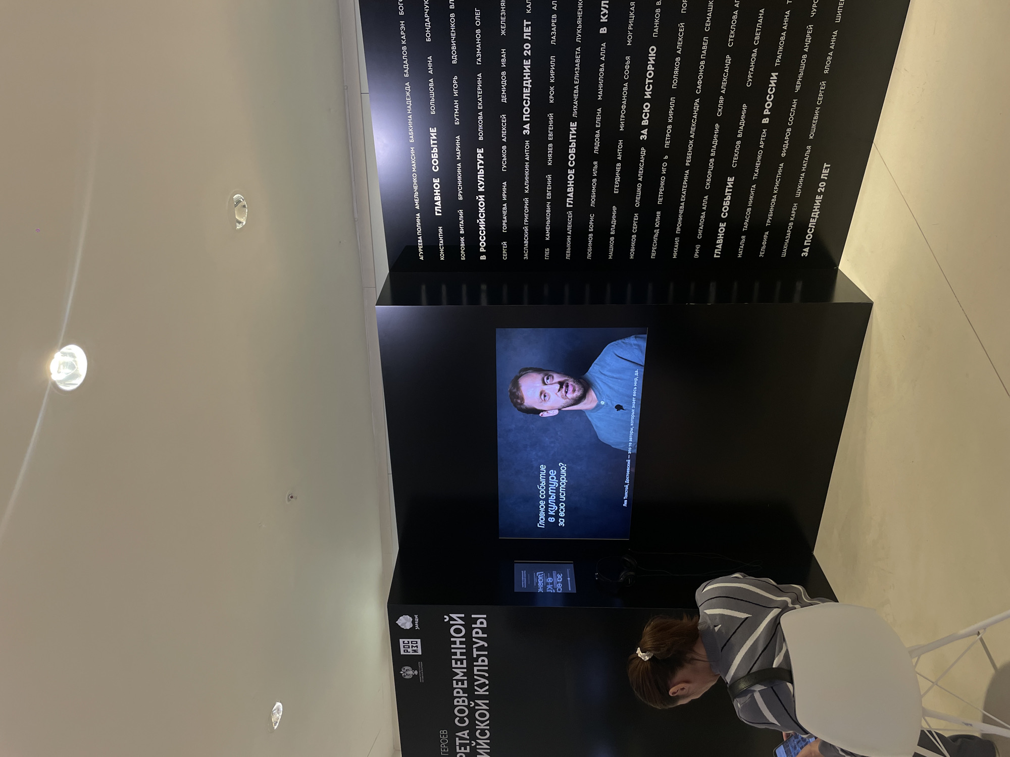 The image depicts an indoor space featuring a black exhibit wall with white text in Russian. The text lists names of individuals under the headings "Главное событие," "В российской культуре," and "За последние 20 лет". A display screen mounted on the wall shows a man speaking, with text about culture and history. A person with striped clothing sits in a white chair, possibly looking at a phone. The location appears to be in Khimki, Russia, based on the museum logo. The exhibit promotes contemporary Russian culture and heroes.
