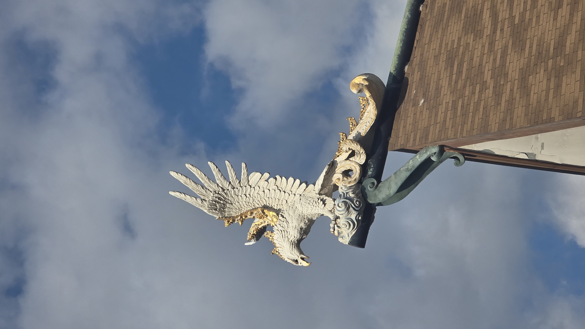 The image features an elaborate, white and gold sculpture of a mythical bird, resembling a Garuda, with its wings spread wide as if captured in dynamic flight or swooping. The bird's body is intricately detailed with feathers, and significant portions of its wings, head, and back are highlighted in a shimmering gold finish. Its beak is open, adding to the sense of motion.
This ornate sculpture is prominently mounted on or integrated into a building's architecture, specifically below the eaves of a roof. The bird is connected to a complex, decorative support structure, which also features gold-colored scrollwork and detailed carvings. Extending from this structure is a gracefully curved, patinated green metal element, possibly a decorative brace or part of a historical drainage system.
Above the decorative elements, a section of the building's roof is visible, covered with uniform, reddish-brown rectangular tiles arranged in a diagonal pattern. The underside of the eaves reveals an off-white surface with some visible cracking.
The entire scene is set against a bright blue sky filled with scattered white, fluffy clouds, indicating daytime. The perspective is from below, looking up at the majestic sculpture as it projects from the building. The intricate design and the likely representation of Garuda are consistent with traditional architectural styles found in Southeast Asia, particularly in Denpasar, Indonesia. No text is visible in the image.