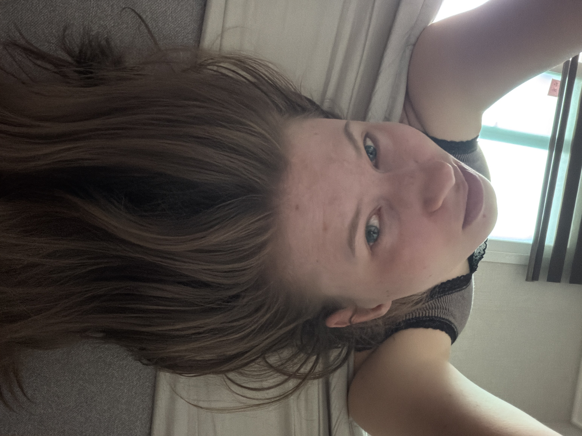 A person is lying on a bed with their head upside down, facing the camera. Their hair is long and brown, falling around their face. They are wearing a striped tank top with black lace trim. The background shows a bed with beige sheets and a glimpse of a window with horizontal blinds. There is a small red sticker with Korean writing on the blinds. The scene appears to be an indoor residential setting.