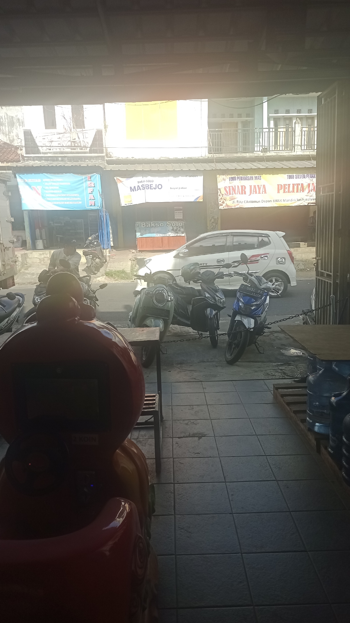 The image depicts a street scene in Karyamandala, Indonesia, viewed from inside a covered area, likely a shop or a covered walkway. In the foreground, a red children's ride-on toy is prominently displayed. Behind it, the street is lined with several parked motorcycles and a white car. A man is visible on one of the motorcycles. Shops are situated along the street, with various signs advertising services and products, including "Bakso Solo Masbejo," "Sinar Jaya Pelita Jaya," and "Toko Perhiasan Mas." The environment suggests a commercial area with moderate activity, indicated by the presence of vehicles and businesses. The lighting suggests it is daytime, with some glare from the sun.