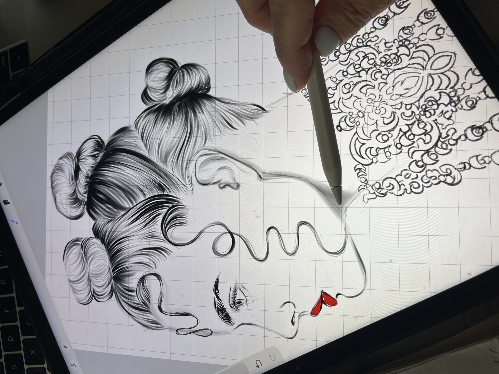 A close-up, overhead shot shows a digital illustration being created on a tablet. The illustration depicts the profile of a woman with elaborate, curled hair styled into two buns. She has striking red lips, detailed eyes, and eyebrows. The background is a white grid, typical of digital art software. A stylus, held by a hand with a light-colored fingernail polish, is actively drawing on the tablet, continuing the intricate linework of what appears to be a decorative garment or accessory below the woman's chin. The tablet is positioned on a surface that shows the edge of a keyboard with the letters S, D, F, G, H, J, K, L, X, C, V, B, N, M visible. The lighting suggests an indoor setting, with no identifiable urban features.