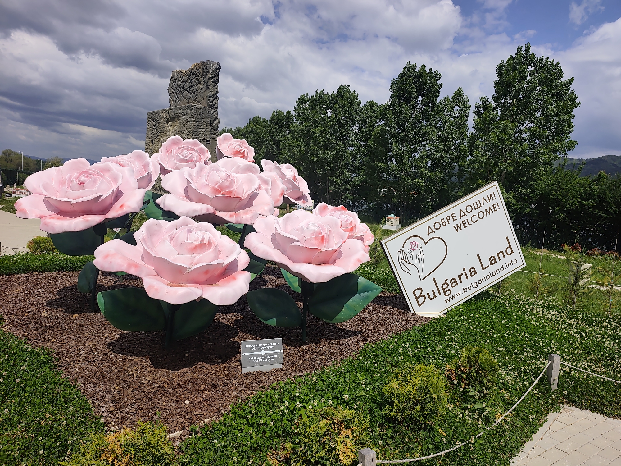 The image shows a display of oversized pink roses in Kazanluk, Bulgaria. In the background is a stone tower, and to the right is a sign that reads "Добре Дошли! Welcome! Bulgaria Land, www.bulgarialand.info". The roses are planted in mulch, and there is greenery around the display. The sky is cloudy.