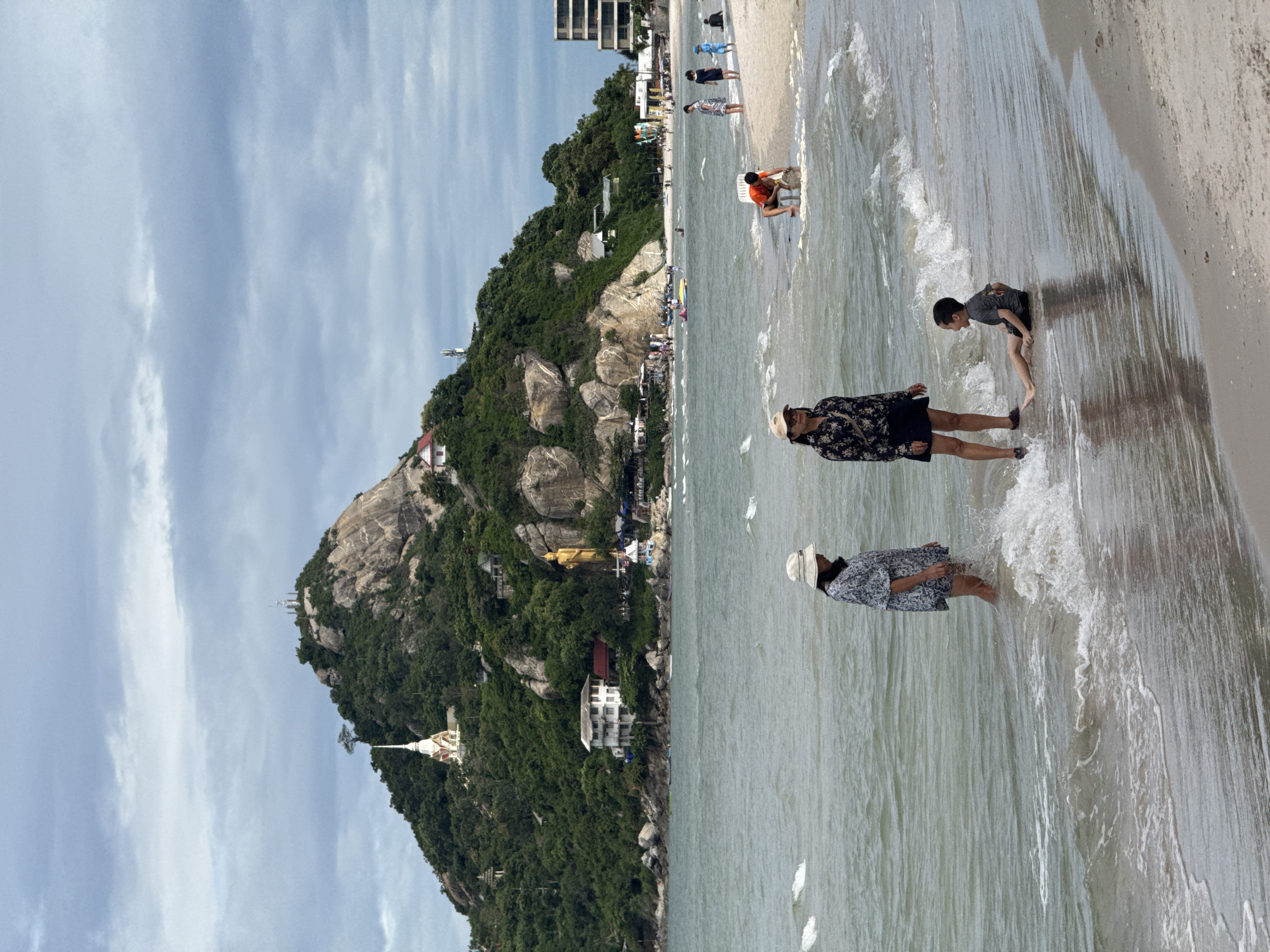In the city of Hua Hin, Thailand, on a cloudy day at the beach, a group of people are enjoying the water. In the foreground, two women and a young boy are in the shallow water near the shore. One woman, wearing a patterned shirt and a wide-brimmed hat, stands partially submerged in the water, facing away from the camera. To her right, a second woman, also wearing a hat and a patterned top and skirt, stands facing the boy. The boy, wearing a dark t-shirt and shorts, is seated on the wet sand in the shallow water, looking up at the woman. Further up the beach, several other individuals are visible. One person in an orange shirt is crouched on the sand. To the right, a group of people, including two children, are standing on the drier sand. The sea is calm with gentle waves lapping the shore. In the background, a lush green hill rises, topped with buildings and a prominent golden Buddha statue. Buildings and a hotel complex are also visible along the coastline, and a few boats are dotted on the water. The sky is overcast with scattered clouds.