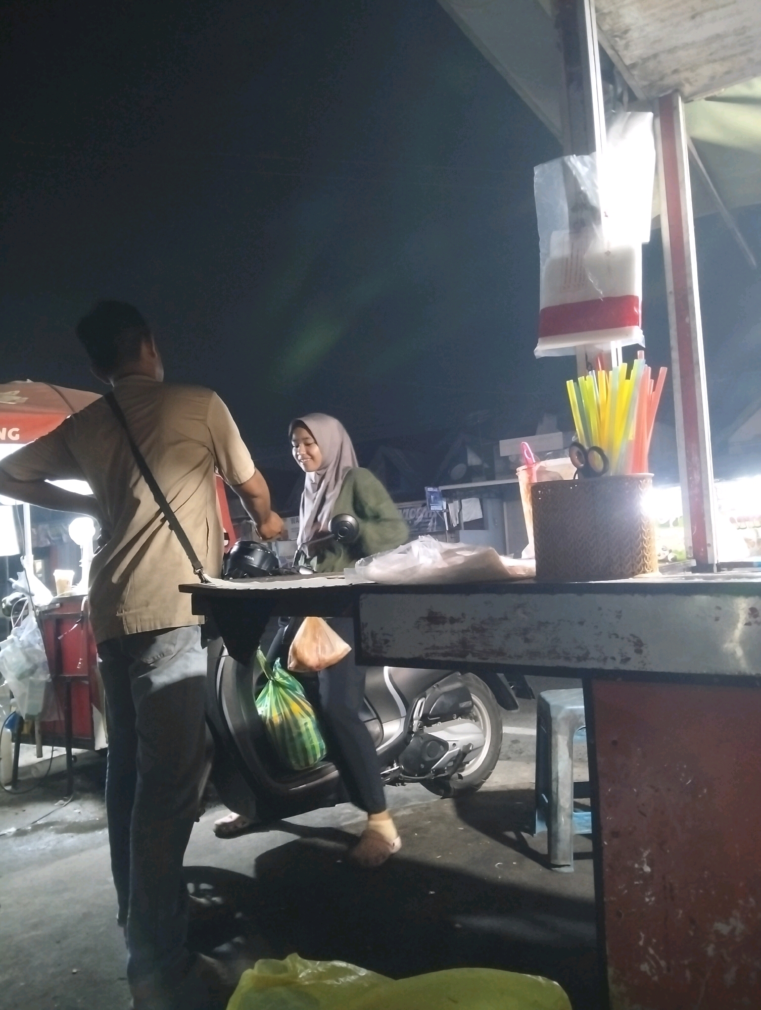 The image depicts a street food stall at night, likely in Palangka Raya, Indonesia. Two individuals are visible: a man wearing a light brown t-shirt and dark pants, and a woman wearing a hijab and a green shirt. The man is standing behind a stall, appearing to interact with the woman. The woman is smiling and looking towards the man, suggesting a friendly interaction.
In the foreground, a counter with various items is visible, including a basket containing colorful straws and scissors. A motorcycle is parked behind the counter, with a green plastic bag hanging on its side. The stall is illuminated by artificial lights, casting shadows on the ground. The overall environment suggests a bustling, open-air market or street scene.
