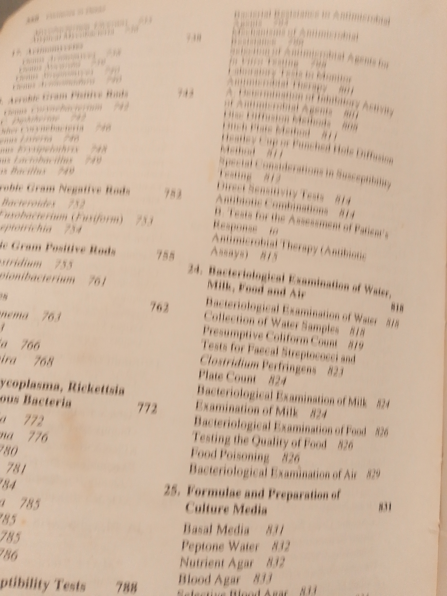 The image is a close-up, slightly blurry, overhead shot of a book's table of contents. The text is printed in columns and lists various scientific or medical topics, likely related to microbiology or bacteriology, with corresponding page numbers. The lighting is artificial, creating some glare and shadows. The background is a plain, light-colored surface, possibly a table. There are no people, objects, or discernible environmental features present besides the book itself. The image is not suitable for determining its location in Jalingo, Nigeria, or any other geographical area, as it contains only text from a book.