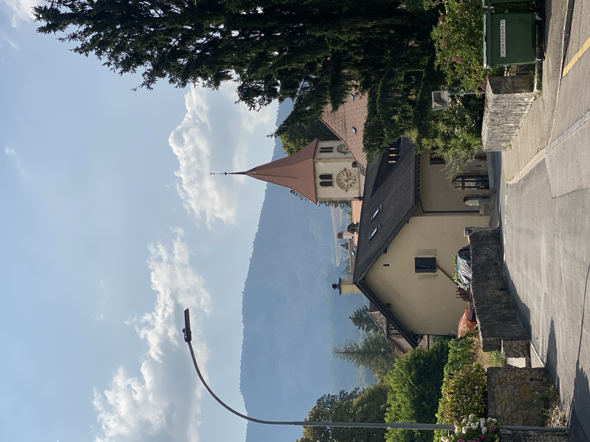 The image shows a street scene in the town of Milvignes, Switzerland, on a sunny day with scattered clouds. A paved road curves to the right, bordered by stone walls. On the left, a lamppost with an LED light fixture arches over the road. Beyond the wall on the left, there are blooming flowers and foliage. On the right, the road continues past a stone wall and two large green recycling bins, one of which has white lettering that reads "CROIX 11-13-15-17".
In the middle ground, several buildings are visible. A light-colored house with a dark roof and a single dark window is prominent. Behind it, a church tower with a red conical roof, a clock face, and a weather vane rises above other buildings. Large evergreen trees are on the right side of the frame, partially obscuring the church tower. In the background, rolling hills covered in trees recede into the distance under a hazy sky. The scene appears to be a quiet, residential area.