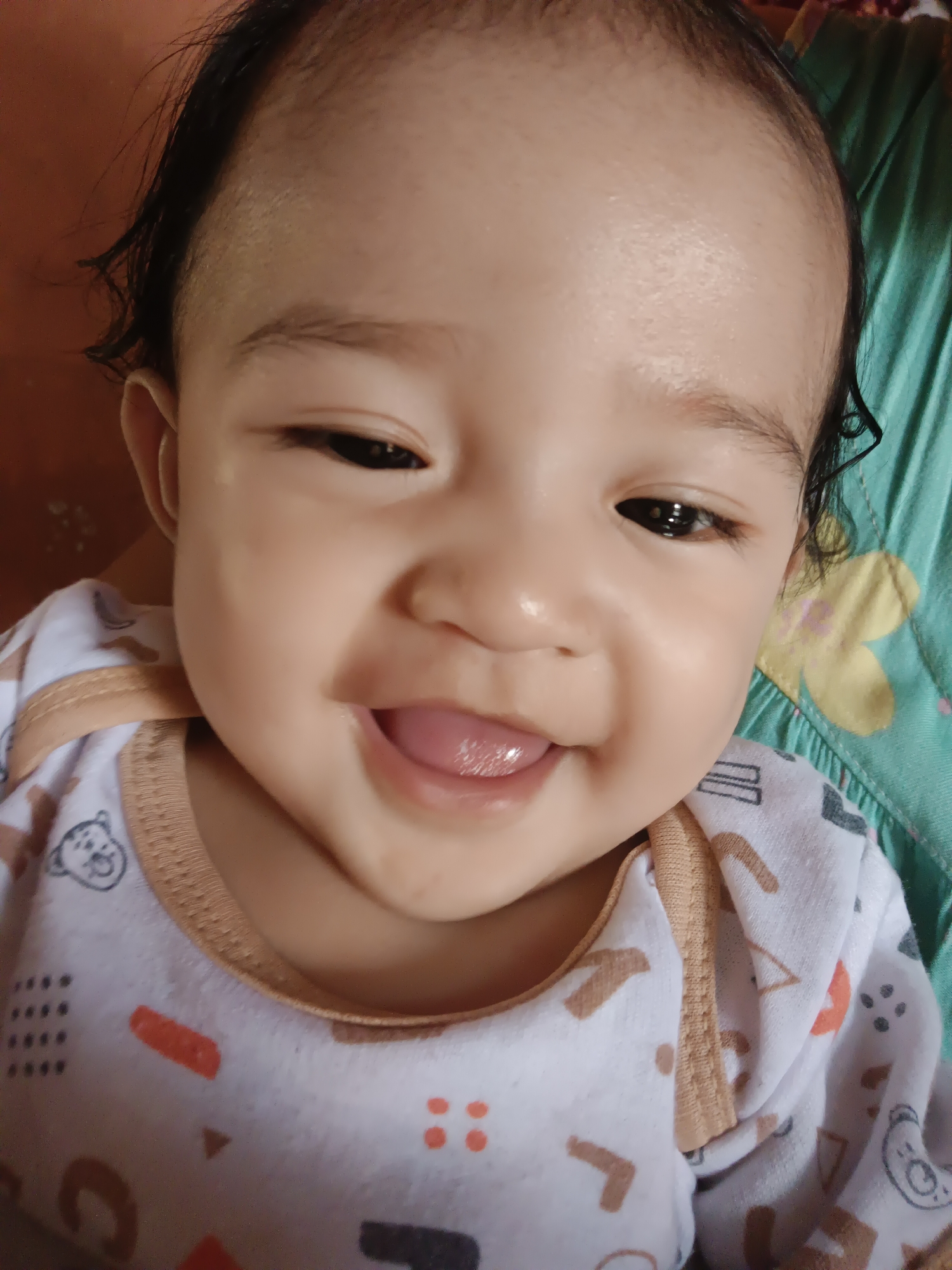 A close-up, head-and-shoulders shot shows a smiling baby's face filling the frame against a blurred background of a brown surface on the left and a teal, patterned fabric on the right. The baby, with dark hair and clear, dark eyes, is wearing a white onesie with a pattern of cartoon bears and geometric shapes. The baby's mouth is open in a wide smile, revealing a pink tongue. The lighting suggests an indoor environment, likely in a home in Kawua, Indonesia, given the context of the request. The image is focused on the baby's joyful expression.